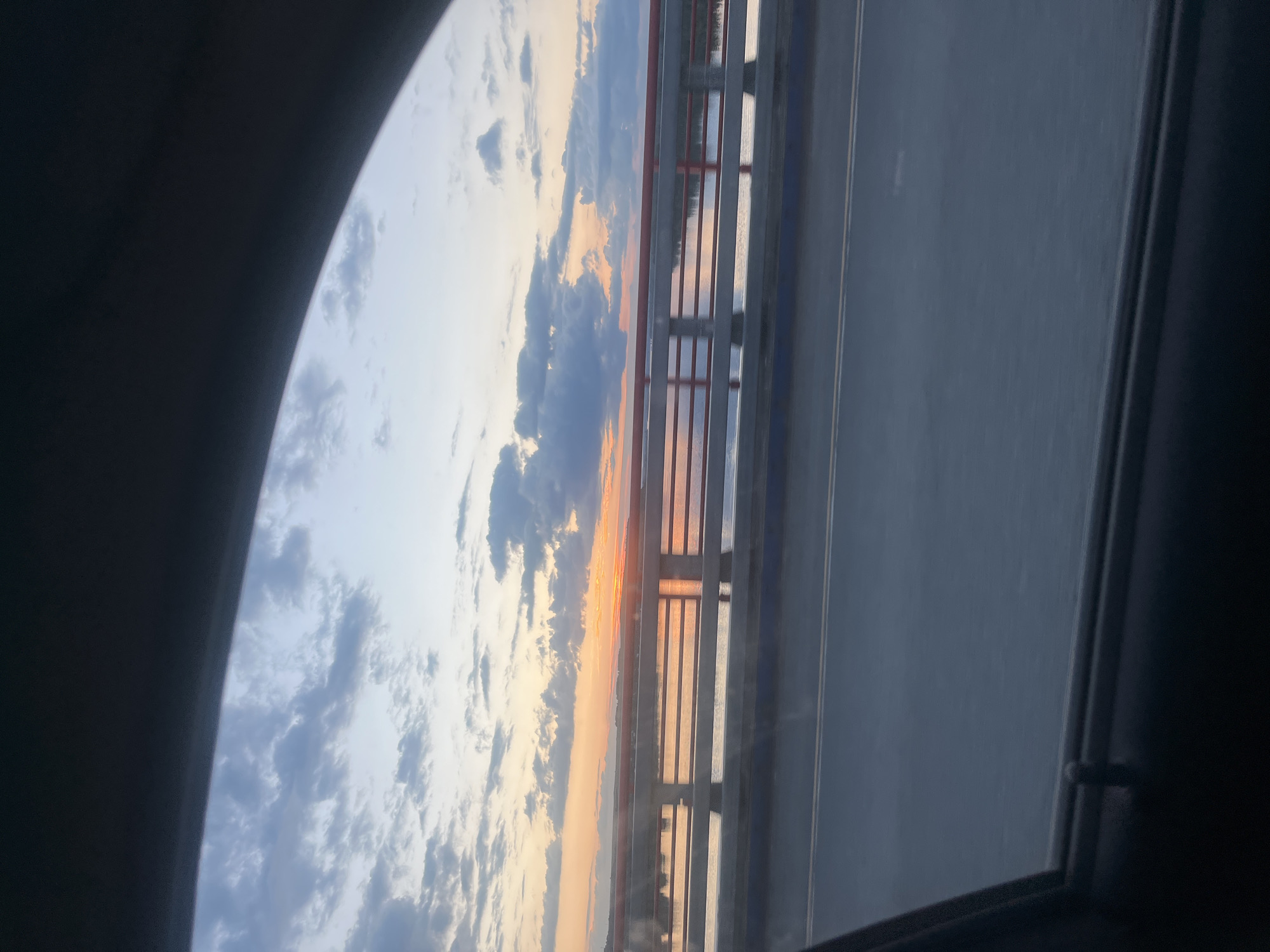 This image captures a view from a vehicle, likely a train, looking out through a window. The dominant scene is a sunset over a body of water, possibly a river, framed by a bridge. The sky is filled with clouds, tinged with the warm hues of orange and pink from the setting sun, transitioning to lighter blues and grays higher up. The bridge structure, with its red railings and vertical supports, is visible on the right side of the frame, casting reflections onto the water below. The water appears calm, mirroring the sky and the bridge.
The foreground is partially obscured by the dark interior of the vehicle, with a curved dark edge suggesting the window frame or a section of the vehicle's interior. On the right side of the image, beyond the bridge, a dark road or pathway is visible, with faint lines suggesting lanes. The overall mood evoked is serene and contemplative, typical of a sunset view during travel. The time of day is clearly sunset, indicated by the colors in the sky. While no people are visible, the perspective suggests they are passengers within the vehicle. There is no visible text in the image.