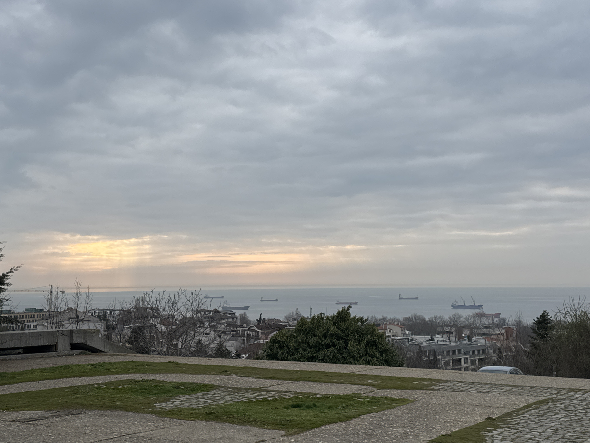 The image shows a cityscape of Varna, Bulgaria, viewed from an elevated position, likely a park or hilltop area overlooking the Black Sea. The foreground features a paved area with patches of grass. Below, there is a view of a densely built urban area with buildings of various heights. Beyond the city, there is a body of water, the Black Sea, with several ships visible on the horizon and closer to shore. The sky is overcast with a diffuse light filtering through the clouds, possibly indicating sunrise or sunset. The overall atmosphere is somewhat muted and grey, typical of an overcast day. There are bare trees mixed among the buildings. There are no people visible in the scene.