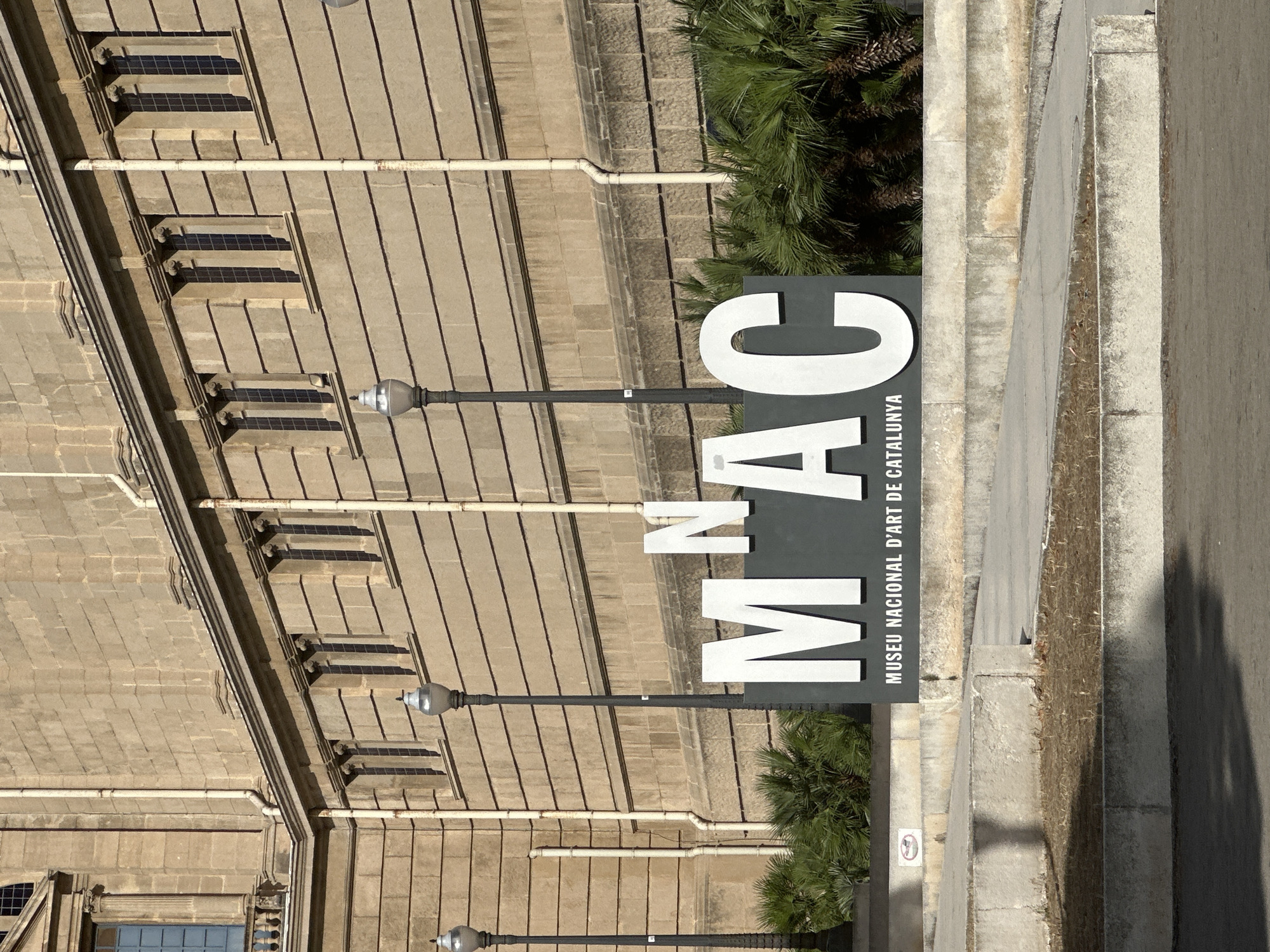 The image shows the facade of a large stone building, likely a museum, with a prominent sign that reads "MNAC MUSEU NACIONAL D'ART DE CATALUNYA". The building has a classical architectural style, featuring rows of arched windows and decorative stonework. In the foreground, there are several large, white letters forming the acronym "MNAC" on a dark background, with the full name of the museum written below it. Palm trees are visible behind the sign. The overall scene appears to be an exterior view of the Museu Nacional d'Art de Catalunya, located in Barcelona, Spain. The lighting suggests it is daytime.