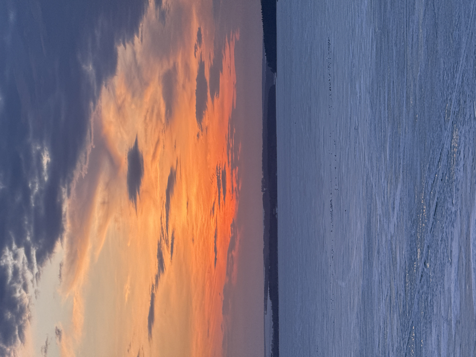 The image displays a landscape divided into two distinct sections. The left side of the image shows a vibrant sunset with hues of orange and yellow streaking through the sky, mixed with dark, cloudy formations. On the right side of the image, a body of water, likely a frozen lake or river, stretches out with visible tracks or lines across its surface. A dark treeline is visible between the sky and the water. The scene is likely taken in Votkinsk, Russia, given the prompt. There is no explicit evidence of people or specific objects. The interaction displayed is that of the setting sun contrasting with the frozen landscape.