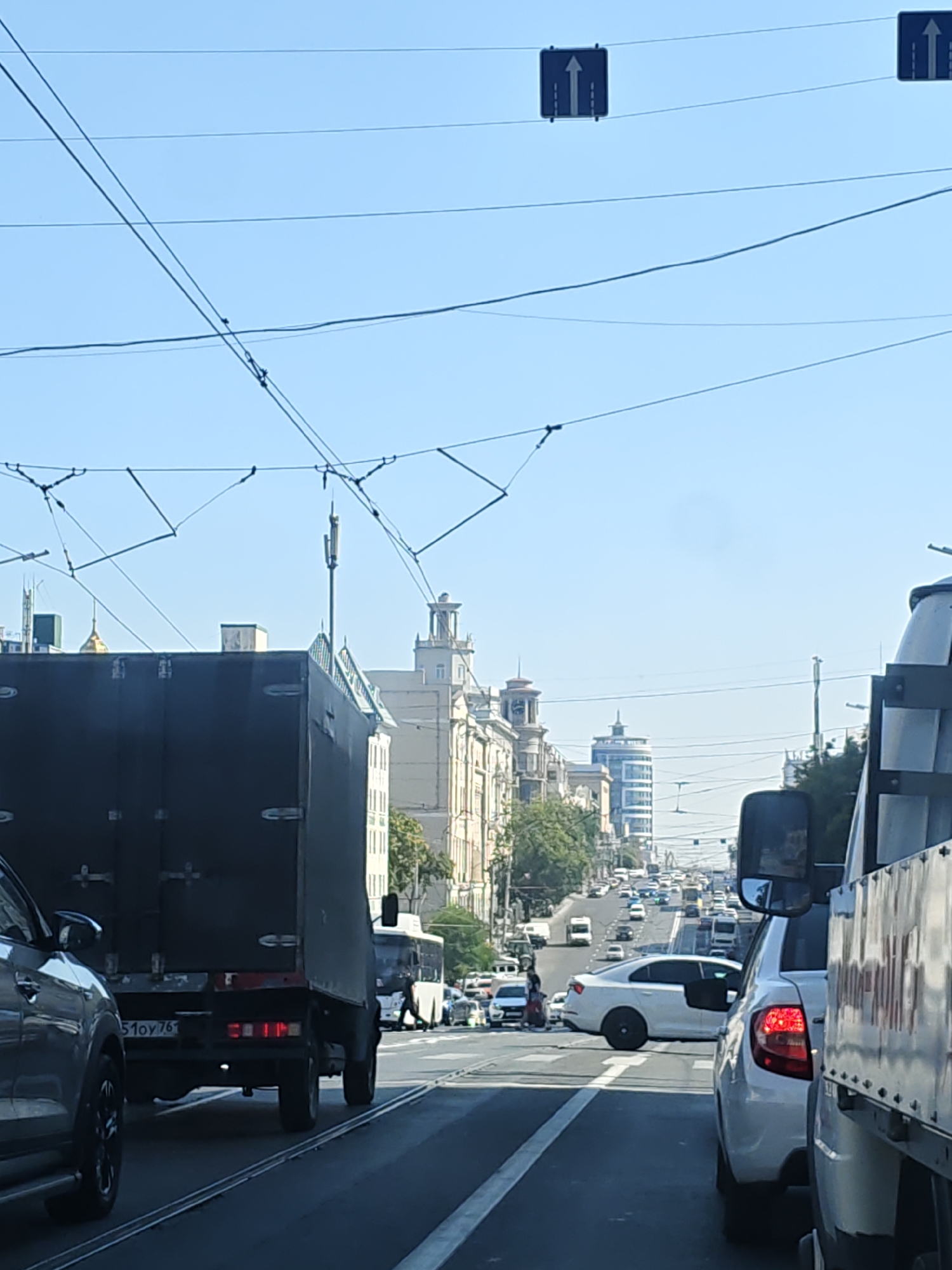 The image shows a daytime view of a busy city street with heavy traffic. In the foreground, a large black truck is visible from the rear, with its license plate reading "51 OY 761". To its left, a silver SUV is partially visible. Further down the road, a white sedan is seen turning, and behind it, a procession of vehicles including cars and buses moves along the street. Several traffic lights are visible, including one directing traffic straight ahead. The street is lined with buildings of various architectural styles, some old and ornate, others modern. Overhead, a complex network of wires, likely for trolleybuses or trams, crisscrosses the sky. The scene suggests a typical urban environment in Rostov-on-Don, Russia, characterized by dense traffic, diverse architecture, and extensive infrastructure for public transportation.