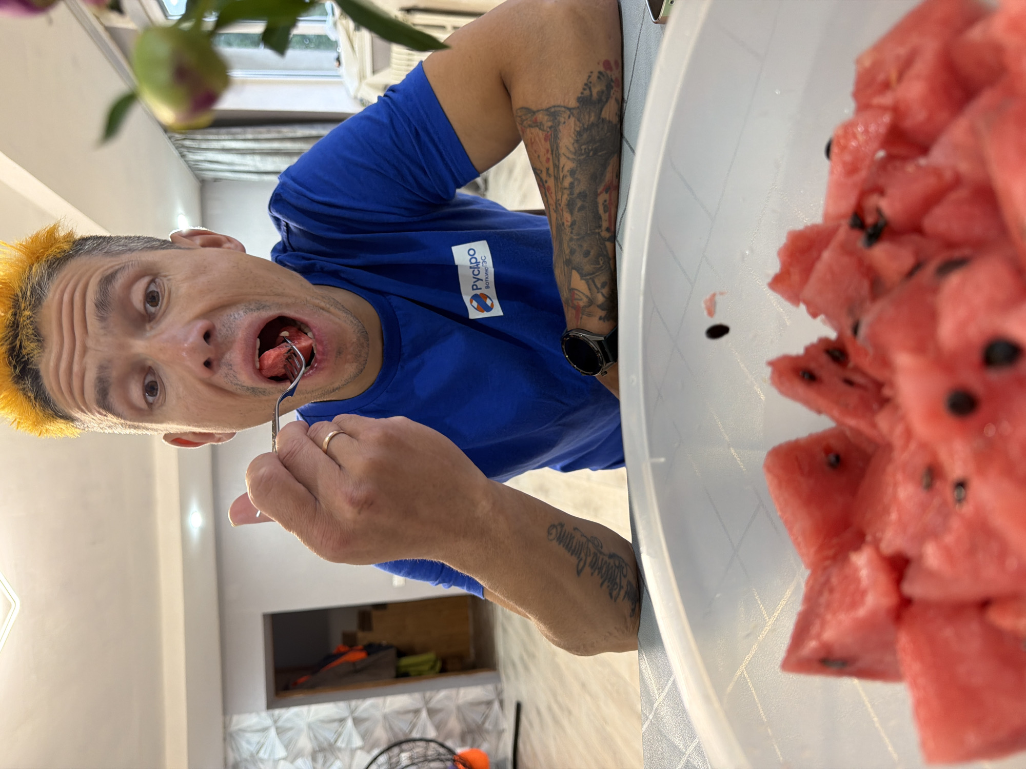 A man with bright yellow hair is in the process of eating a slice of watermelon with a fork. He is wearing a blue t-shirt with a logo that includes a white rectangle with a red and blue stripe and the Cyrillic text "РусИЗО". He also has a large tattoo on his arm. He appears to be in a casual indoor setting, possibly a kitchen or dining area. The room is brightly lit, suggesting it is daytime. A large bowl of watermelon slices is visible in the foreground. In the background, there are glimpses of furniture and what appears to be a fan. The image has been taken from a low angle, looking up at the man.