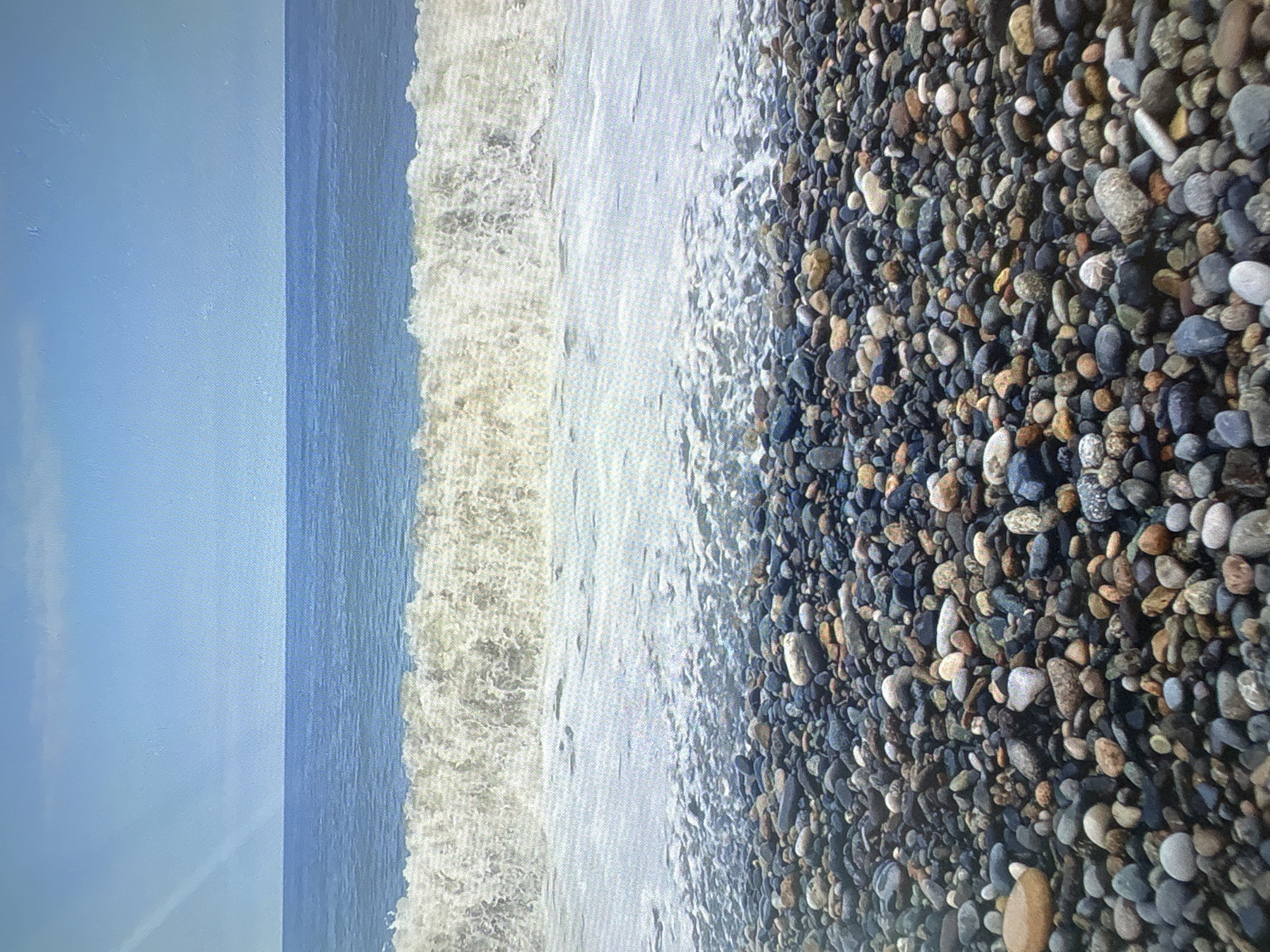 The image depicts a pebbled beach in Batumi, Georgia. The foreground is covered with a dense arrangement of various colored and sized pebbles. A wave is breaking, with white foam washing over some of the stones and receding into the sea. The sea is a deep blue, meeting a clear blue sky with a few wispy clouds. The overall scene is a serene coastal landscape.