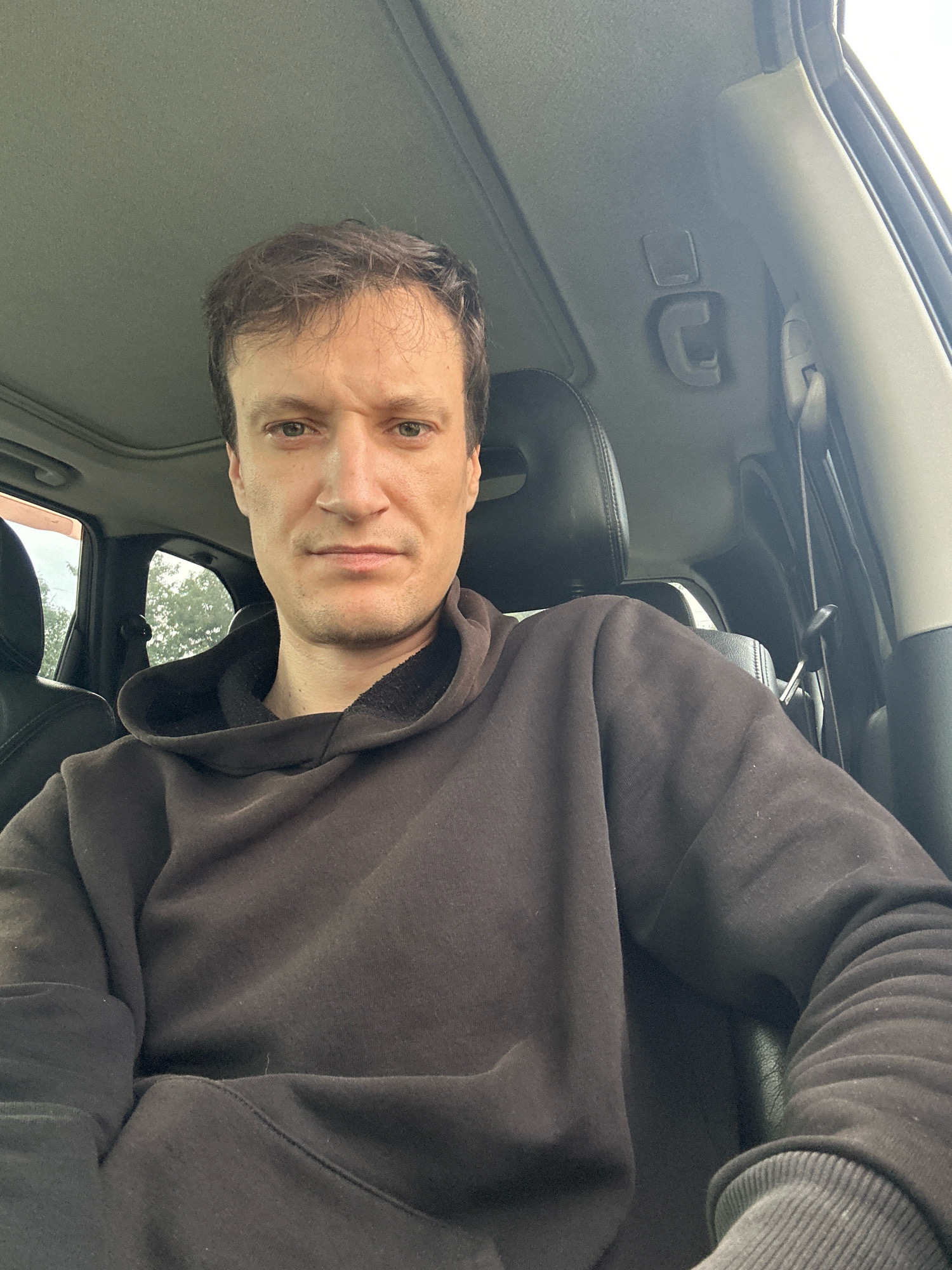 A man is sitting in the driver's seat of a car, looking at the camera. He has short, brown hair, and is wearing a dark brown hooded sweatshirt. The car's interior is visible, including the headrest of the passenger seat and a portion of the sunroof. The background outside the car appears to be trees and foliage, suggesting a rural or suburban setting.