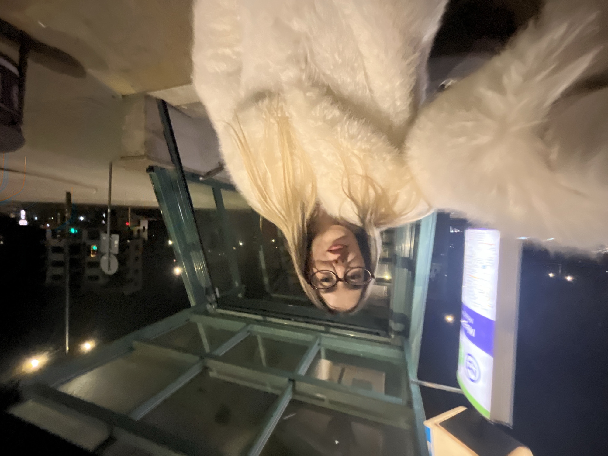 The image shows a woman with blonde hair and glasses, wearing a white, furry coat, seemingly taking a selfie while upside down in the city of Sofia, Bulgaria.
She is positioned within a glass-enclosed structure, potentially a bus stop or similar urban fixture. The structure has a metal frame and visible panes of glass. Outside of the structure, the background suggests an urban environment at night, with streetlights and indistinct buildings visible. A sign with blue and white colors is also visible next to the glass structure. The scene's overall lighting appears to be dim, with artificial lights providing the primary source of illumination.