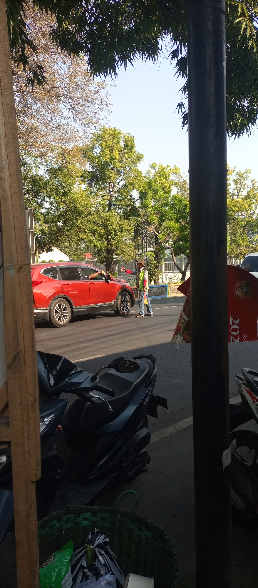 The image depicts a street scene in Jatirejo, Indonesia. A red SUV is parked on the right side of the road, facing left. A person wearing a reflective vest and a hat stands next to the driver's side of the SUV, appearing to interact with someone inside the vehicle. In the foreground, several dark-colored motorcycles are parked, with a green woven basket filled with trash situated in front of them. Trees with lush green foliage line the background of the street, with a clear blue sky overhead. A dark pole is positioned on the right side of the image, partially obscuring the view of the background. The overall impression is that of a typical urban street environment in Indonesia, likely during daylight hours.