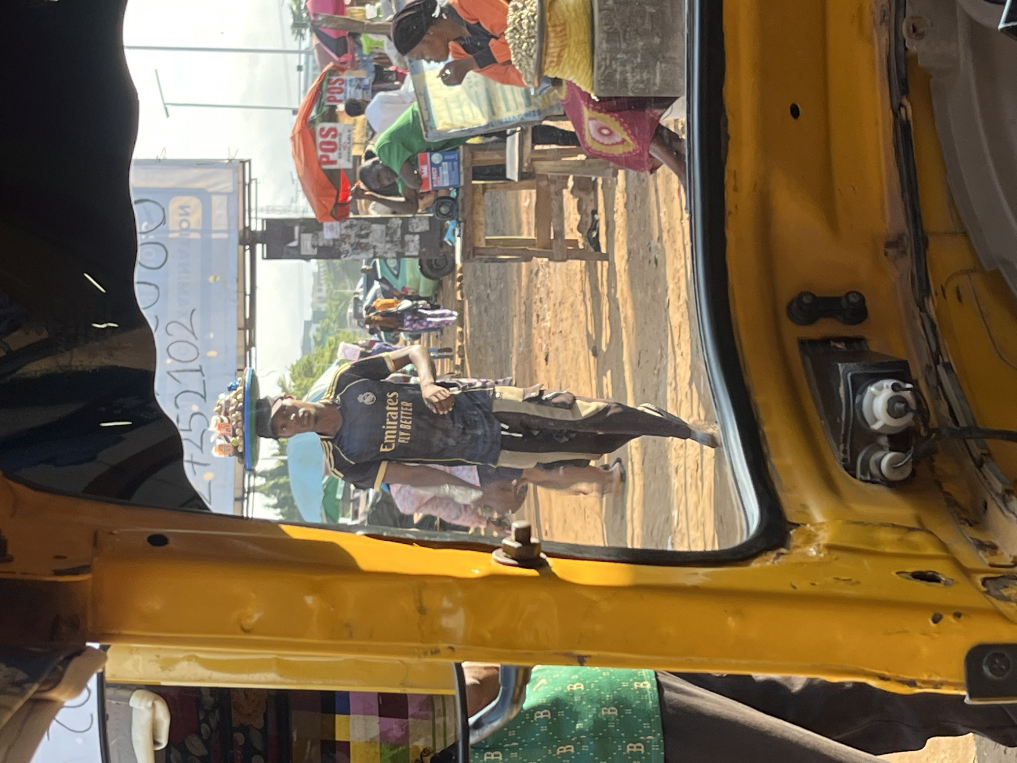 A man wearing a black and gold athletic jersey walks while carrying a tray of food on his head in what appears to be an outdoor market setting in Abuja, Nigeria. The man is walking from left to right in the frame, his body angled slightly away from the camera. The food on the tray is colorful and appears to be a variety of snacks or small dishes. He is wearing dark pants with lighter stripes down the sides.
The scene is framed by the yellow interior of a vehicle, likely a taxi or rickshaw, suggesting the image was taken from inside. The foreground shows parts of the vehicle's interior, including yellow painted panels and some electrical components.
In the background, other people are visible, some walking and others interacting near stalls. There are colorful umbrellas and signs, typical of a market environment. Some of these signs appear to be for "POS" services, indicating financial transaction points. The ground is unpaved, suggesting a dusty or dirt road. Sunlight is casting shadows, indicating it is daytime. The overall atmosphere appears to be bustling and active.