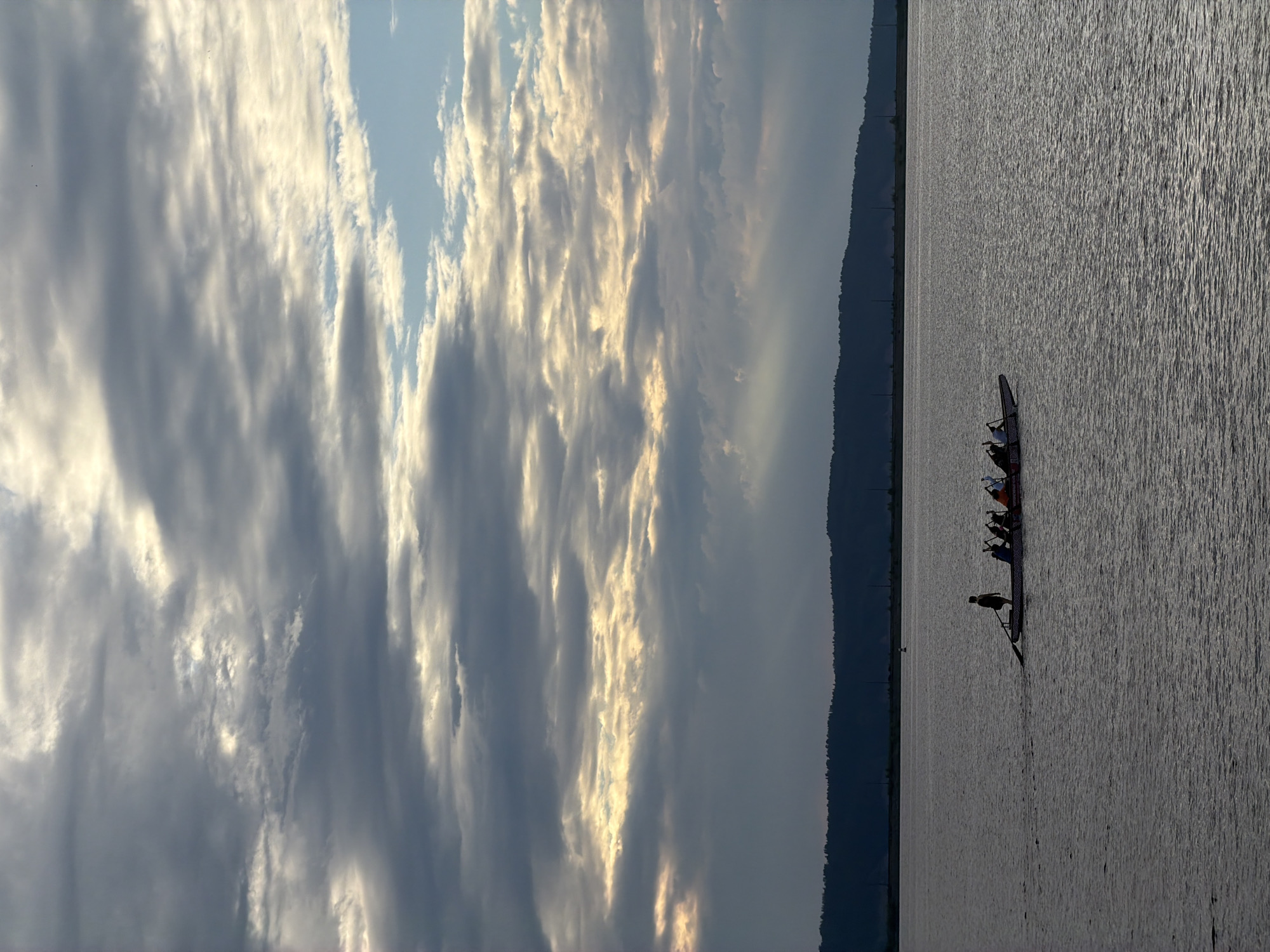 The image depicts a serene, wide-angle view of a calm body of water, likely a river or lake, under a dramatic sky. The water's surface is rippled, reflecting the subdued light of what appears to be either early morning or late evening, given the soft, diffused lighting. A long, narrow boat, possibly a dragon boat or a similar racing craft, is the primary object on the water. It is occupied by several people, who are silhouetted against the bright water. One individual in the rear of the boat appears to be steering or in a central position, while others are seated further forward, suggesting they are rowing or preparing to.
The sky is filled with a dynamic arrangement of clouds. Some are dark and heavy, while others are illuminated by sunlight filtering through them, creating streaks of light and shadow across the expanse. This celestial display creates a sense of vastness and natural beauty.
In the background, a dark, treeline forms a distinct horizon separating the water and sky. This suggests a natural landscape, possibly a forested area along the coast or riverbank. The overall mood is peaceful and contemplative, with a focus on the interplay of light, water, and human activity within a natural setting. There is no visible text in the image. The location context of Chaikovsky, Russia, suggests a setting in that region.