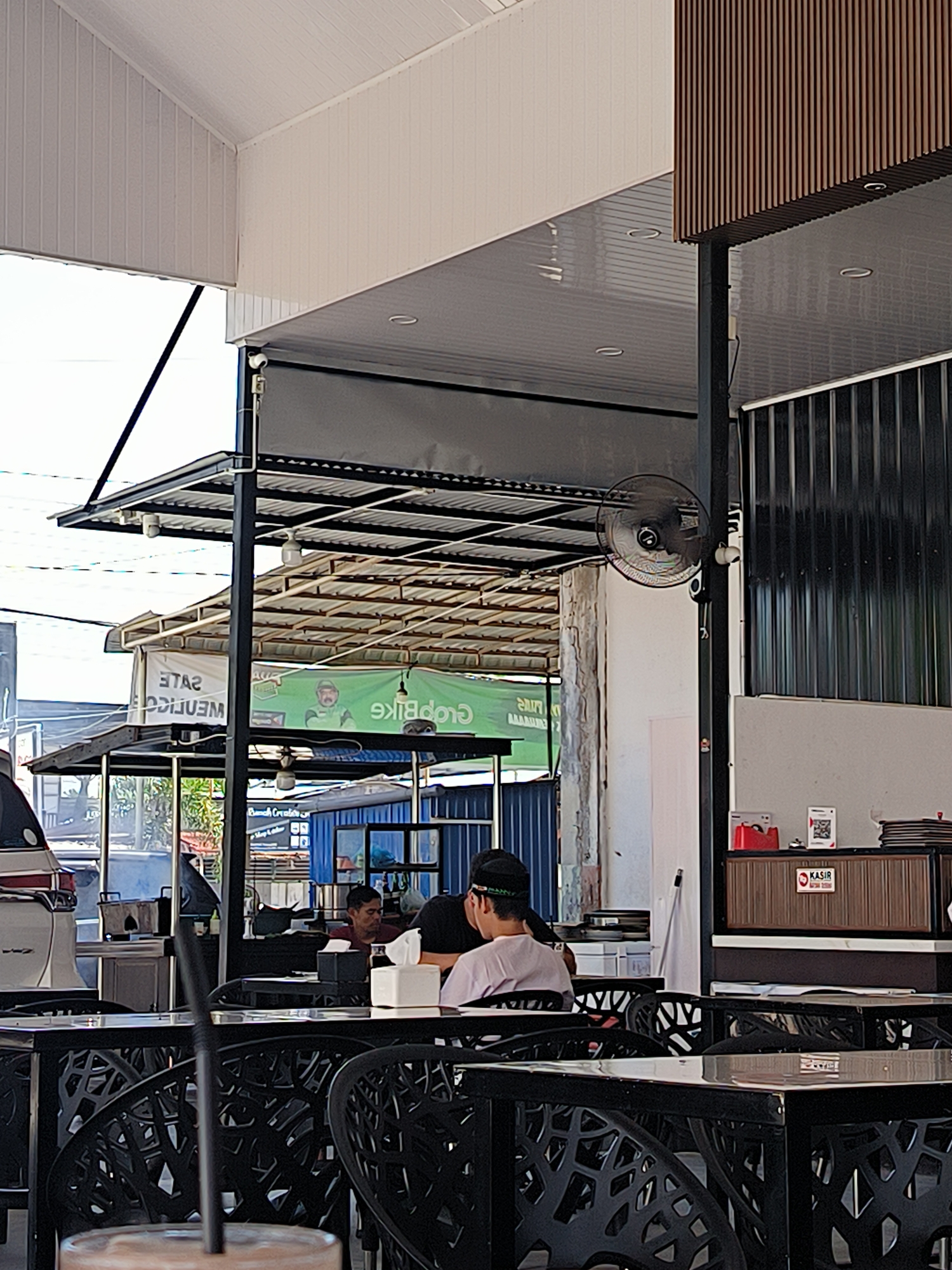 The image captures an outdoor seating area, likely a cafe or restaurant, in Lhokseumawe, Indonesia. Several individuals are seated at black metal tables with ornate chairs. A car is partially visible in the background, along with commercial signs, including one prominently advertising "Crop Bike". A wall-mounted fan is visible. The roofing is a mix of structures and canvas, creating a partially covered space. A person wearing a cap is visible. The weather is bright and sunny.