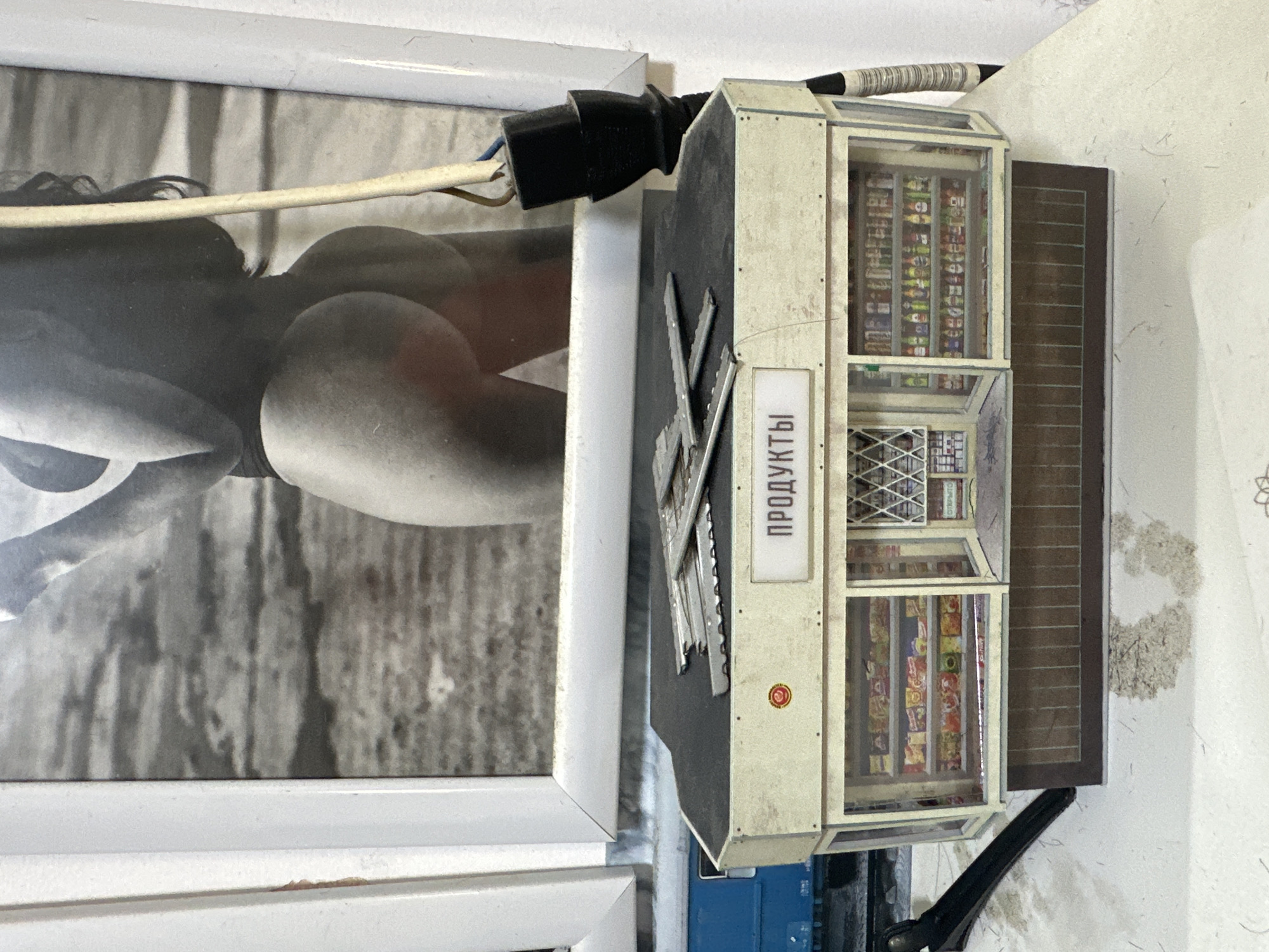 The image shows a collage of seemingly unrelated items. A black and white photograph of a woman's rear is framed in white. Next to the photograph is a miniature model of a "Products" (ПРОДУКТЫ in Russian) store vending machine, with a power cord plugged into the side. The vending machine appears to be filled with miniature food products. The setting appears to be indoors, with a white wall in the background. There is no information to place this image in Tel Aviv, Israel.