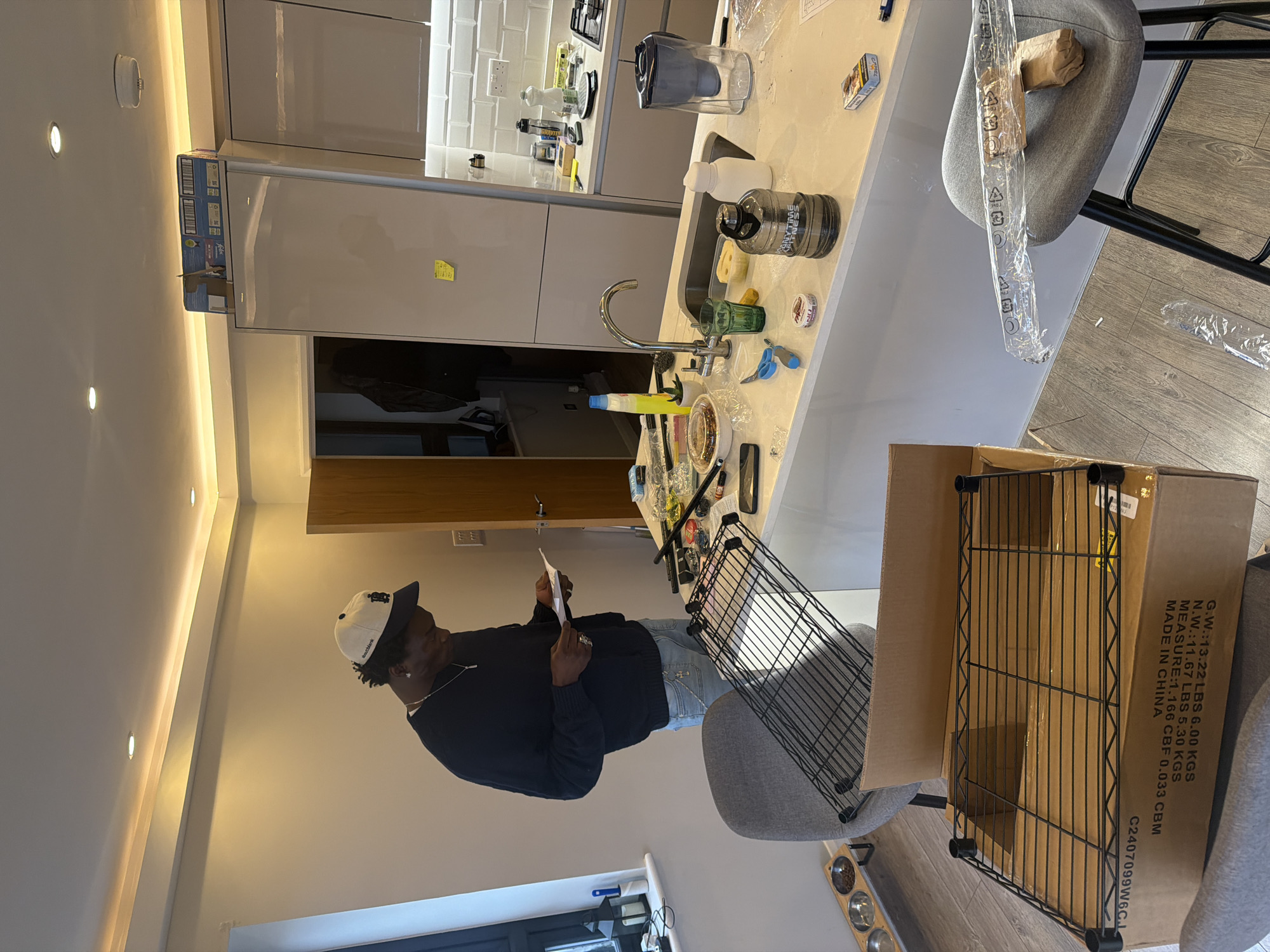 The image depicts an indoor domestic scene, specifically a modern kitchen and living area, in London, United Kingdom.
A male individual, with dark skin, wearing a dark blue long-sleeved sweater, light blue ripped jeans, and a white baseball cap with a dark brim and a "B" logo, stands in the center-left. He is holding and reading a white sheet of paper, likely assembly instructions. A silver chain with a cross pendant is visible around his neck, and he wears a ring on his right hand.
The primary subject of assembly is a black wire shelving unit. One partially assembled shelf rests on an open cardboard box in the foreground, labeled "MADE IN CHINA" with weight specifications in both imperial (LBS, CBF) and metric (KGS, CBM). Another black wire shelf is placed on a grey upholstered bar stool positioned to the left of the man. A second identical bar stool, on the right, holds clear plastic packaging material and a brown paper-wrapped item. The flooring is light-colored wood laminate.
The background features a contemporary kitchen. A white kitchen island with an integrated sink and a chrome swan-neck tap occupies the midground. The countertop is cluttered with various items related to the assembly process and daily use, including a yellow spray bottle, a small potted succulent, scissors, screwdrivers, a mobile phone, a takeaway container, a green glass, a large grey "ANYTIME FITNESS" water bottle, a white water bottle, a clear plastic water filter jug, and a pack of "Camel" cigarettes.
Beyond the island, a white tiled backsplash (subway tiles) is visible, featuring two white double power outlets with switched rectangular pins, characteristic of UK electrical standards. Above the backsplash are light-colored, glossy kitchen cabinets and a large, silver-grey refrigerator/freezer with a yellow sticky note affixed to its door. A gas hob with a kettle is visible on the far right counter.
To the far left, a window sill displays decorative lanterns and a wooden stand holding two stainless steel pet food bowls. An open doorway with a dark brown frame leads to another room, where a dark jacket hangs. The ceiling is white, featuring recessed spotlights and continuous perimeter LED strip lighting, along with a white smoke detector above the refrigerator.