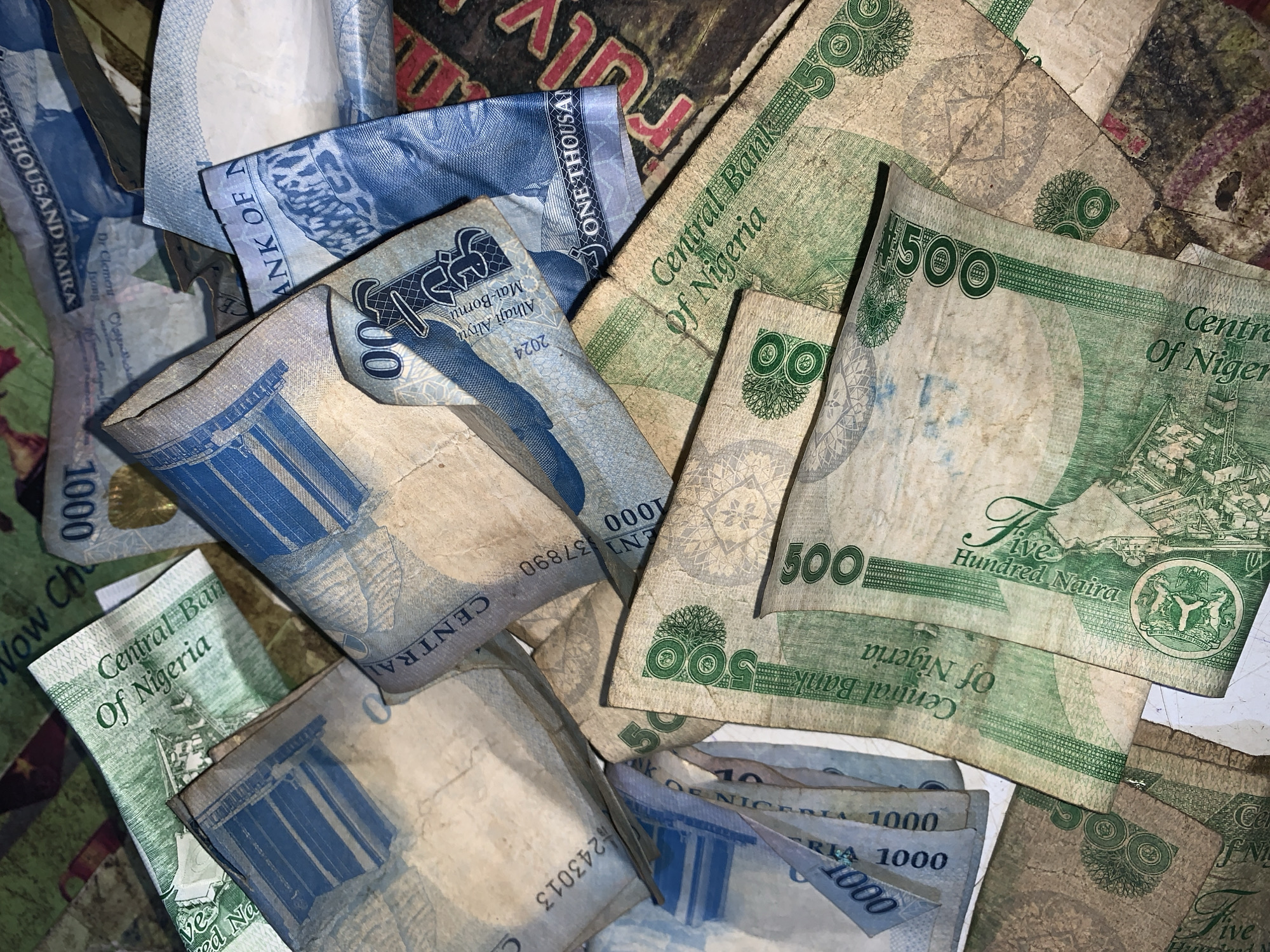 The image shows a disordered pile of Nigerian Naira banknotes. There are both 1000 Naira (blue) and 500 Naira (green) denominations visible. The bills appear to be used and somewhat crumpled. Text on the bills indicates "Central Bank of Nigeria." There is no evidence of a location in the city of New Stadium, Nigeria, and there are no people present.