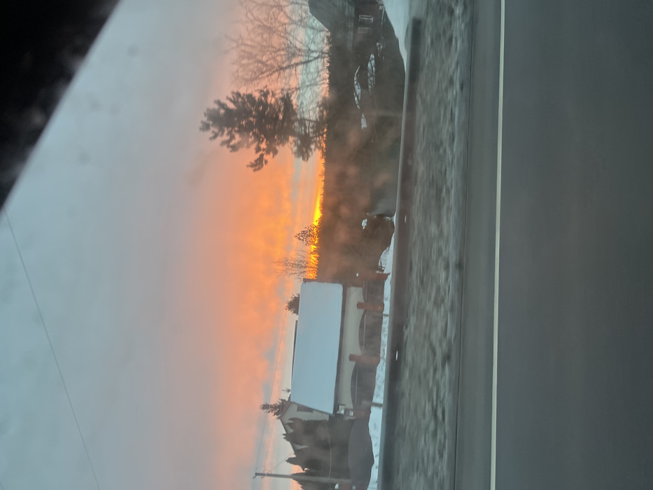The scene depicts a winter landscape at sunrise or sunset in the city of Ширяево, Russia, observed from within a moving vehicle. The sky dominates the upper portion of the image, displaying a gradient from a pale blue-grey at the top to vibrant orange and yellow hues near the horizon, indicative of low sun angle. The sun itself is not directly visible but its light creates a strong glow.
In the midground, from left to right, there is a two-story house with a prominent white, snow-covered gable roof and light-colored walls featuring dark arched openings and brick-like pillars. Utility poles with overhead lines are visible to the left of the house. Bare deciduous trees and dark evergreen trees are scattered across the midground, with their silhouettes partially obscuring the bright horizon. The ground beneath the trees and structures is covered in snow.
The foreground consists of a dark asphalt road with a faint white lane line visible near the bottom center. Immediately above the road, a grey metal barrier (guardrail) runs horizontally. Beyond this barrier, a snow-covered embankment with sparse, dry vegetation extends towards the buildings and trees. The entire scene is viewed through a window, evident by a dark diagonal obstruction in the upper right corner and minor smudges or reflections across the surface of the image. No people or discernible actions are visible.