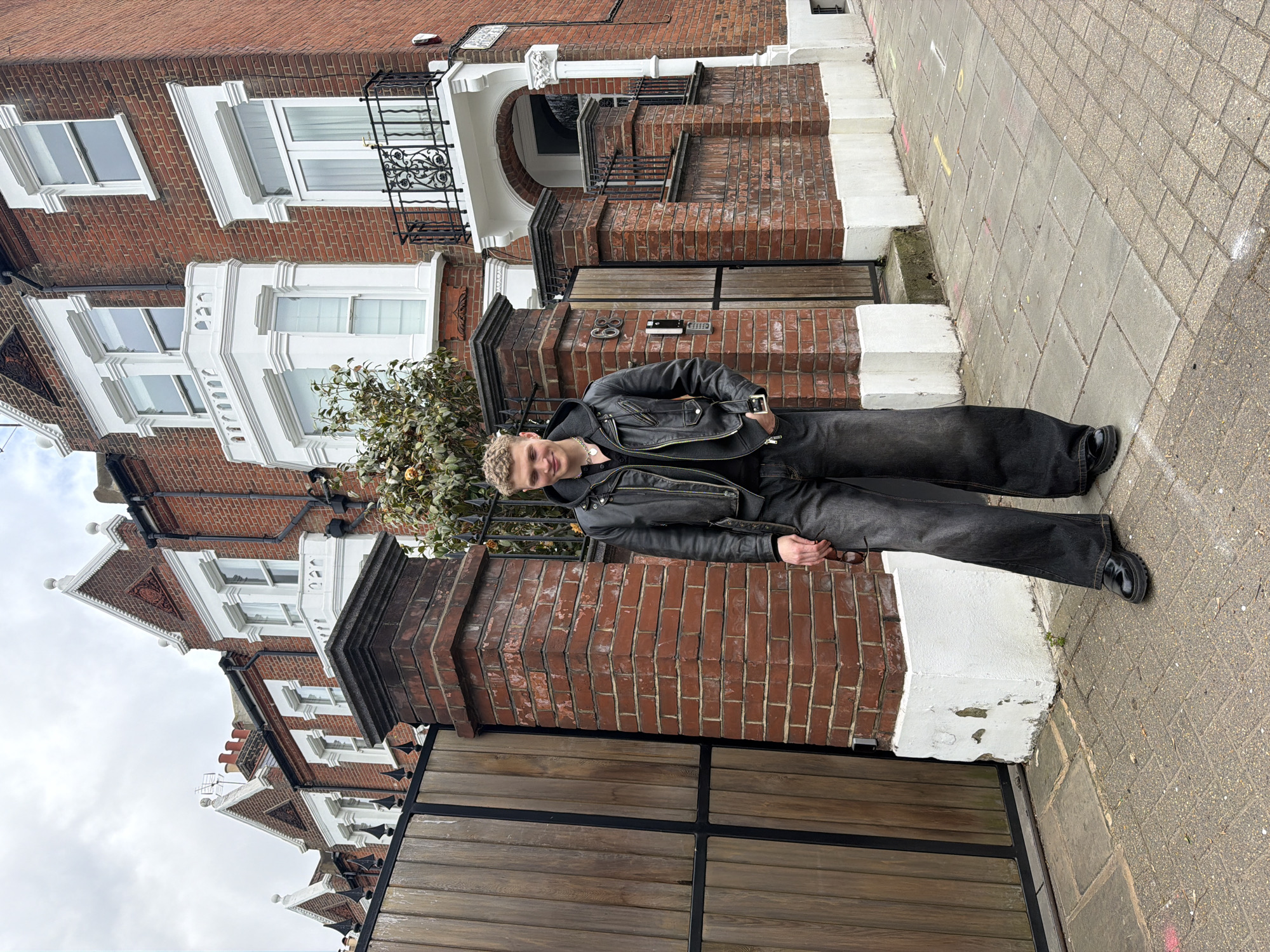 A young man stands in front of a red brick building in London, UK. He wears a black leather jacket, black pants, and black shoes. The building has white window frames and a black wrought iron balcony. The street is paved with gray brick. Number 68 is visible on the building.