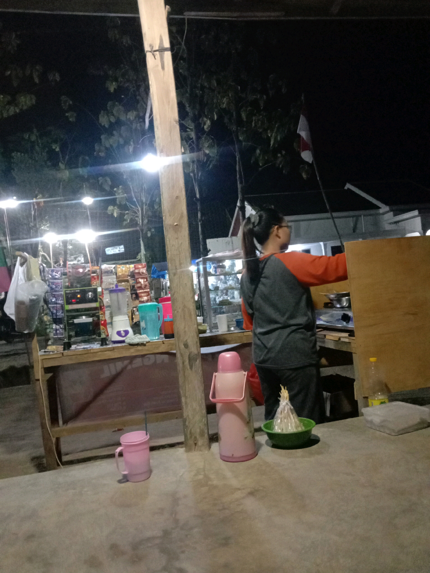 At night, a young woman with her hair in a ponytail and wearing glasses stands behind a stall in Utama Jaya, Mataram, Indonesia. She is wearing a grey and orange long-sleeved shirt and dark pants. The stall is stocked with various items, including bags of snacks, a blender, and a pink thermos. In front of the stall, a pink mug sits on a concrete surface. The area is lit by string lights, and a flag is visible in the background.