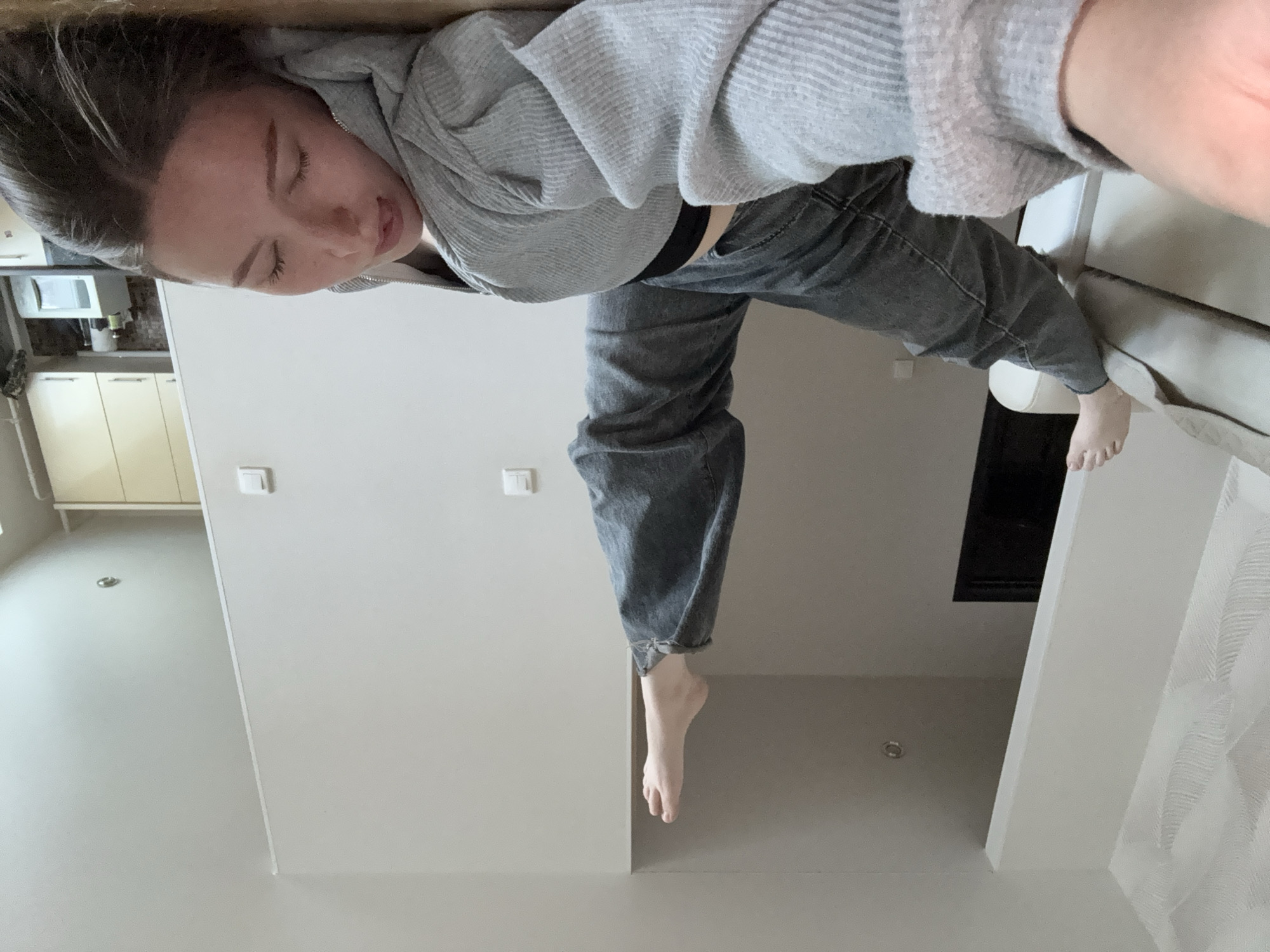 A woman in a gray hoodie and gray jeans is photographed from a low angle. She is lying on a surface, possibly a couch or bed, with one leg dangling toward the ceiling and the other resting on the couch. The location is possibly an apartment, with a view of kitchen cabinets in the background. The scene is in Chaikovsky, Russia.