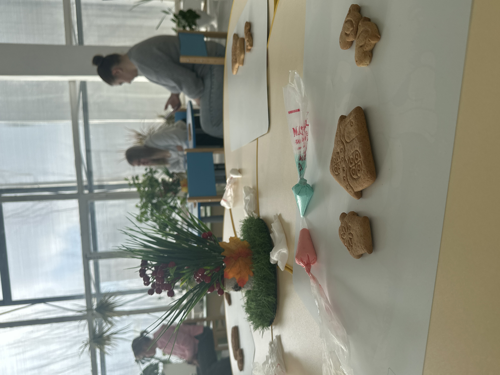 Here is a forensic description of the image:
The image appears to be taken indoors, possibly in a community center or activity room. A long table is covered with a white sheet, upon which gingerbread cookies are scattered. Several icing bags, filled with colored icing, are placed alongside the cookies. A floral arrangement with green leaves, red berries, and an orange leaf is centrally located on the table. Two women are seen in the background, presumably decorating cookies. The room has large windows. The location in the city is Unknown.