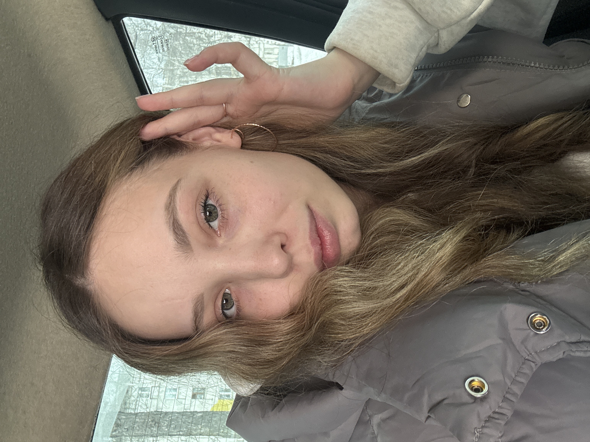 A person is indoors, possibly inside a vehicle, against a window with an indiscernible view that includes trees and buildings. The person has long, wavy light brown hair, fair skin, and light-colored eyes. They are wearing a gray hooded jacket with metallic snap buttons. The person is wearing a gold hoop earring on their right ear. The location in the city of Izhevsk, Russia, cannot be confirmed from this image.