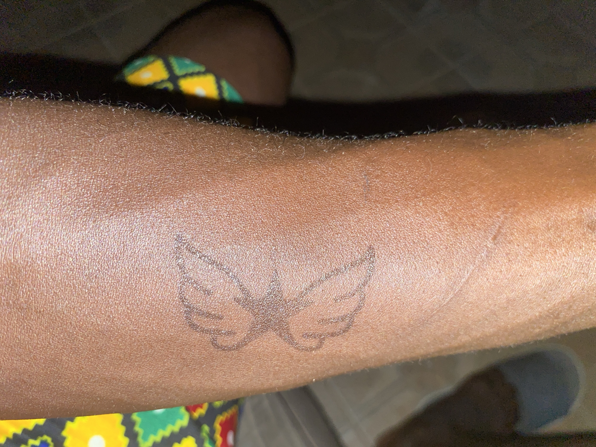 A close-up image shows a person's dark-skinned arm. A tattoo depicting a pair of wings with a star between them is visible on the arm. Part of a patterned garment is visible near the arm. There is no information that would indicate the location of Barden Yana, Nigeria.
