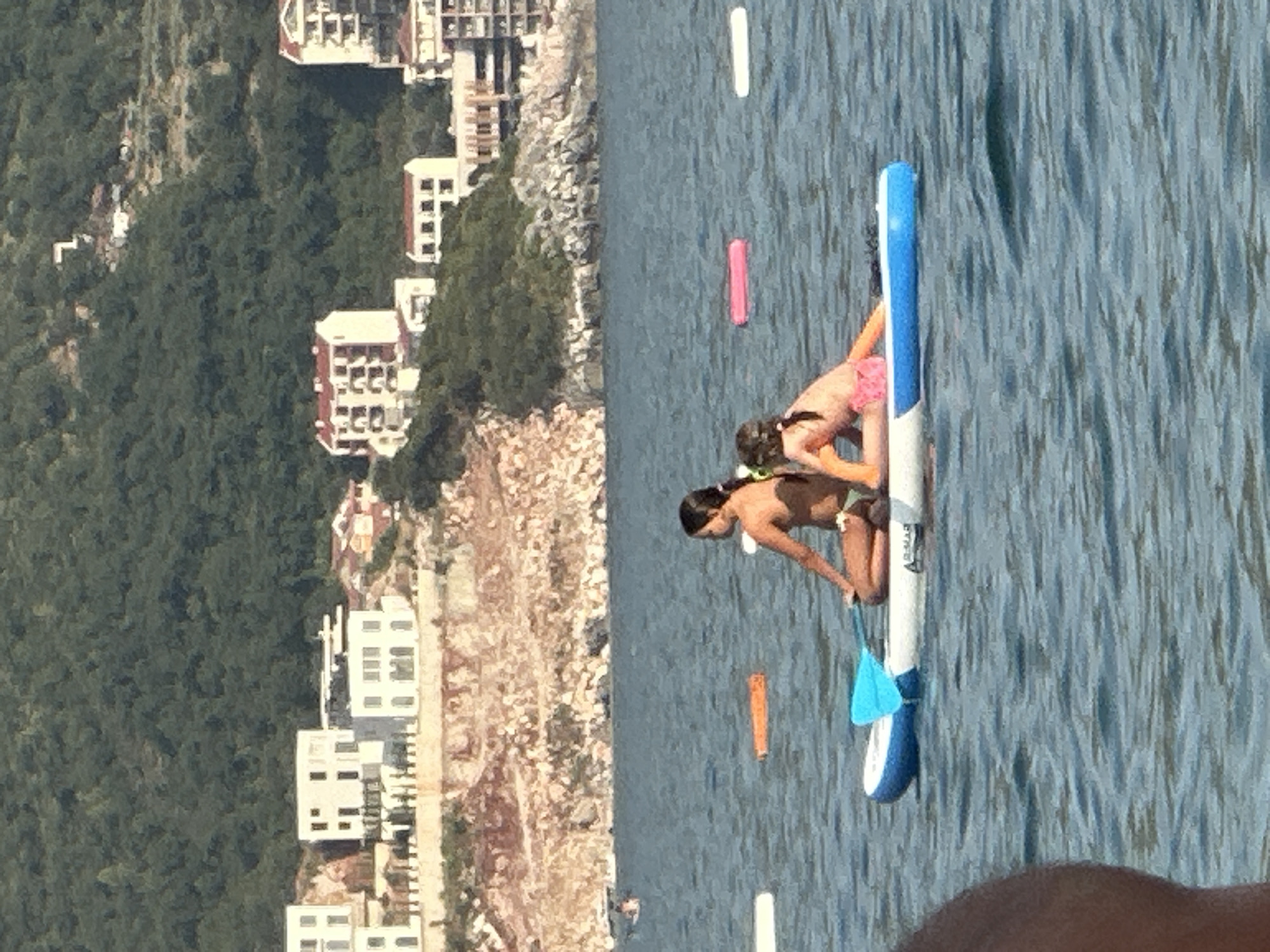 The image depicts a sunny day in Rafailovići, Montenegro, with the scene split between a lush, forested hillside dotted with buildings and a calm, blue body of water. On the water, two individuals are engaged in paddleboarding. One person, wearing pink bikini bottoms, is kneeling on a paddleboard, appearing to balance or prepare to paddle. Another person, further ahead on the same board, is also in a kneeling position, looking down at the water. A bright blue and white paddle is attached to the paddleboard.
In the background, several white buildings with red-tiled roofs are visible, nestled into the green hillside. Some of these buildings appear to be hotels or apartments, with multiple floors and windows. The rocky terrain is also evident, adding to the natural beauty of the location.
Scattered on the water are inflatable pool floats, one pink and one orange, suggesting a recreational atmosphere. The water is relatively still, reflecting the bright sunlight. The overall impression is one of leisure and outdoor activity on a clear, warm day. There is no visible text in the image.