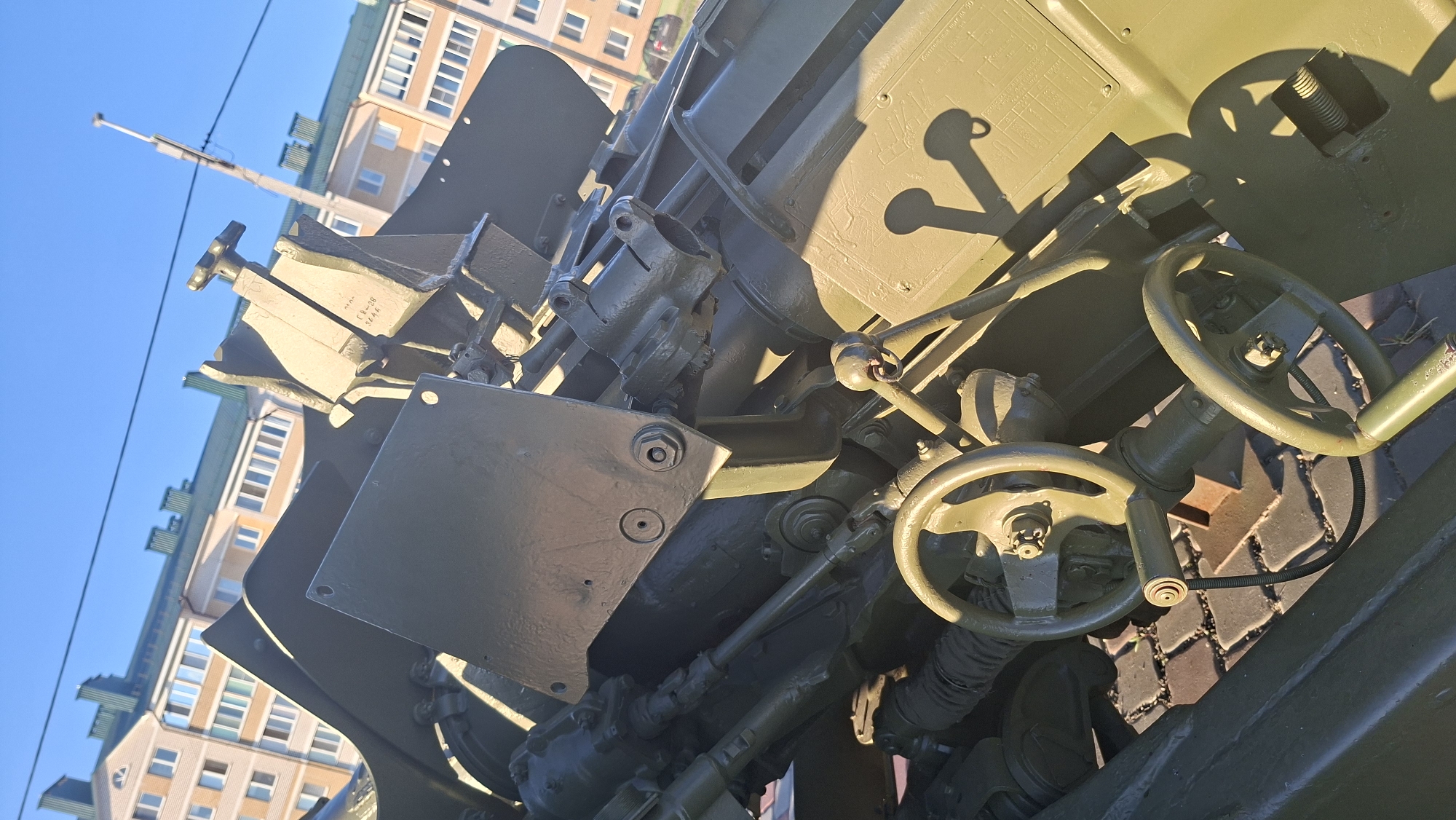 The image shows a close-up of a green military-grade artillery piece, likely a howitzer, positioned outdoors. The weapon is angled, with its intricate mechanical components and aiming mechanisms visible. Two handwheels are prominent in the foreground, suggesting manual adjustment controls. The artillery piece is set against a backdrop of a blue sky and a residential building, which appears to be multi-story with light-colored facade and dark green roofing. The building's architecture suggests it is located in a city. The ground beneath the artillery piece is paved with cobblestones. The lighting indicates it is daytime, with sunlight casting distinct shadows. The presence of the artillery piece in a civilian setting suggests it might be part of a memorial, a museum exhibit, or a public display. Based on its location in the city of Smaliavichy, Belarus, it is plausible that this artillery piece is a historical monument or part of a military-themed display.