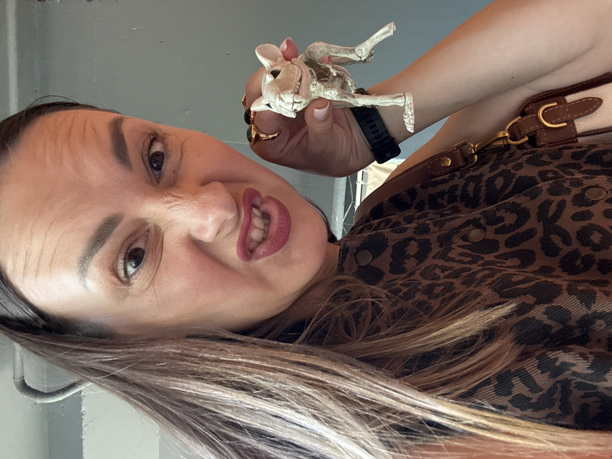 A woman with brown hair pulled back is making a silly face, scrunching up her nose and baring her teeth, while holding a small, tan-colored skeleton figurine of what appears to be a dog or rat in her right hand. She is wearing a brown top with a leopard print pattern and a gold ring on her right ring finger. A dark watch is visible on her left wrist. A portion of a brown and white purse is visible at the bottom of the frame, attached to her by a strap. The background is a plain, light gray wall. There is no indication that this image was taken in Sochi, Russia.