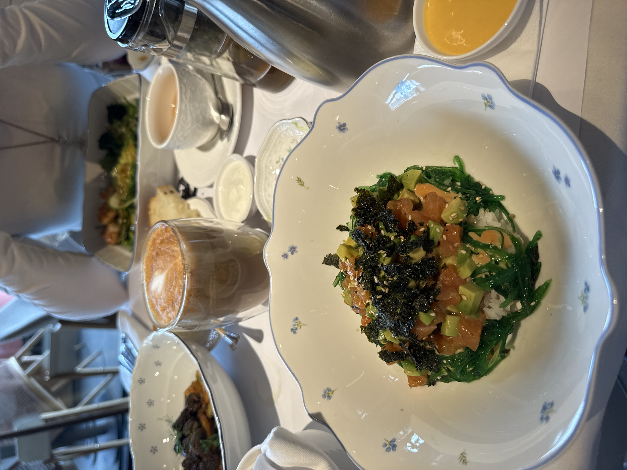 This image presents an overhead view of an elegantly set table laden with a variety of food and drinks, suggesting a luxurious dining experience, possibly breakfast or brunch, in an indoor setting during the daytime.
The focal point is a white, scalloped-edge ceramic bowl featuring delicate blue floral patterns. Inside, there's a vibrant dish resembling a poke bowl, built on a base of white rice. It is generously topped with bright green wakame (seaweed salad), diced orange-pink salmon or tuna, and cubes of green avocado. The dish is artfully garnished with shredded dark green nori seaweed and a sprinkle of white sesame seeds.
Arranged around this central dish are several other items. To the left, a clear glass mug holds a creamy coffee beverage, likely a latte or cappuccino, distinguished by a clear heart-shaped latte art design on its foamy surface. Further back, a white bowl contains a dark, savory-looking dish, possibly a meat or mushroom-based entree. A classic white teacup with an intricate relief pattern and its matching saucer are also visible. Smaller white dishes hold a creamy white sauce (perhaps sour cream or yogurt) and a piece of bread or pastry. In the upper right, a small oval bowl contains a bright yellow sauce or puree. A tall, reflective metal pitcher or thermos is also present in the background.
A person wearing a white long-sleeved top is partially visible on the far left, suggesting they are seated at the table. The overall presentation is refined and meticulous, indicative of a high-quality establishment. The background is softly blurred but suggests an interior space with some structural elements. No specific text or branding is discernible on any of the visible items. Given the context, this sophisticated meal is taking place in Moscow, Russia.