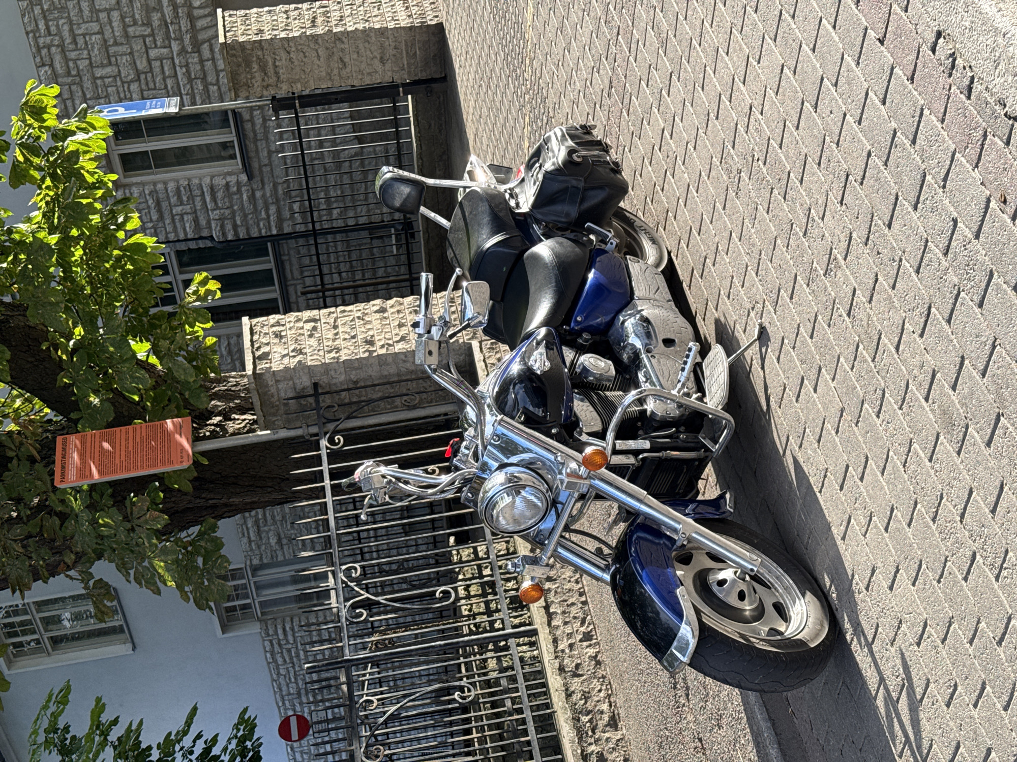 A blue and black cruiser motorcycle is parked on a herringbone patterned brick sidewalk in Tallinn, Estonia. The motorcycle is facing to the left of the frame, with its front wheel closer to the viewer. The motorcycle has chrome accents, a black leather seat and saddlebags, and a tall backrest. In the background, a light-colored stone building with windows is visible, along with a black metal fence and a tree with green leaves. An orange parking sign is attached to the tree. A red and white circular "no entry" sign is also visible on the fence. The scene is illuminated by sunlight, casting shadows on the sidewalk.