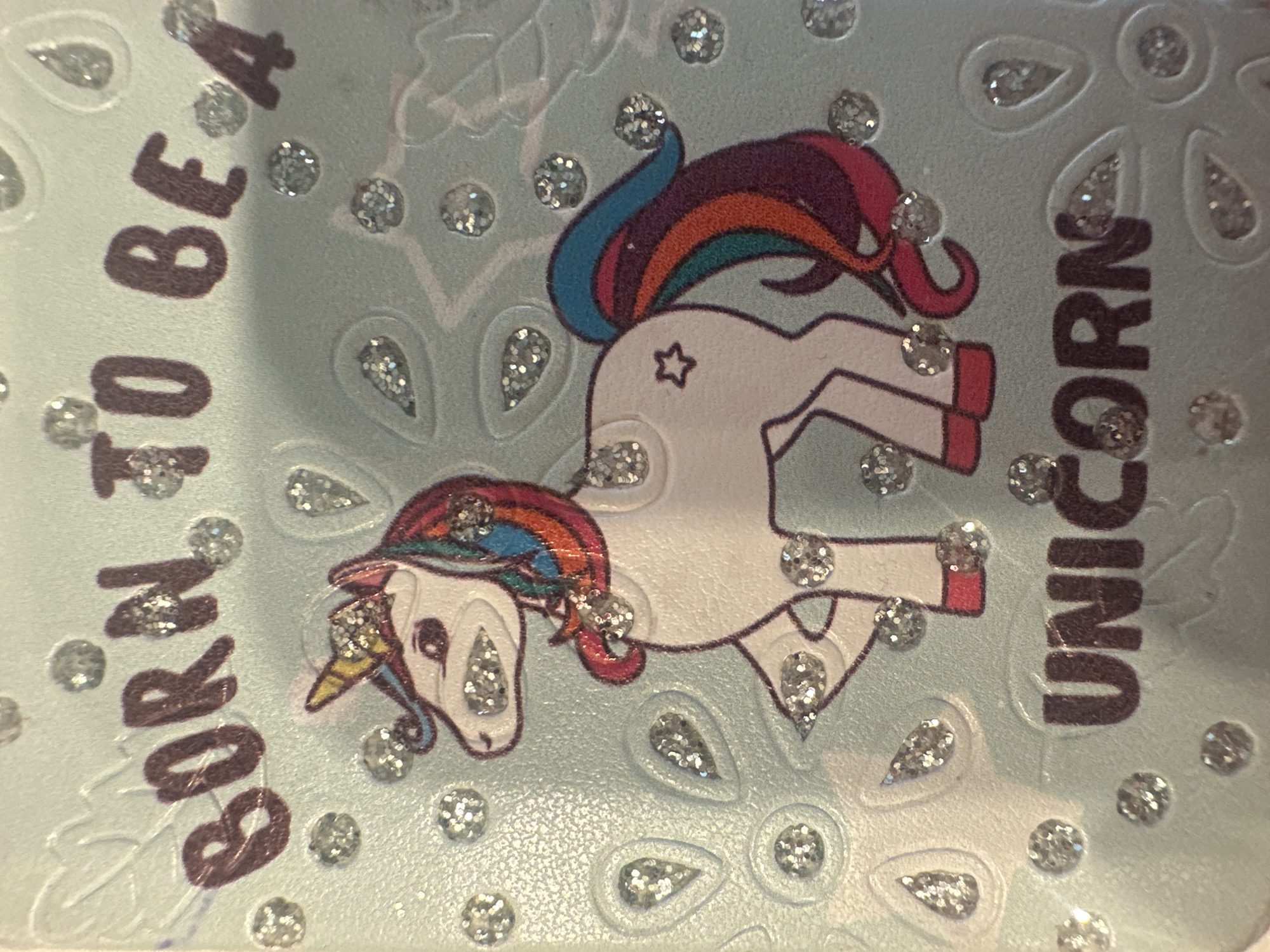 The media file is a close-up, overhead shot of a light blue, textured surface adorned with glittery embellishments and a drawing of a unicorn. The unicorn is depicted in profile, with a rainbow-colored mane and tail, a star on its hindquarters, and glitter accents on its horn, eyes, and hooves. The overall design suggests a whimsical and childlike theme.
Visible text, oriented vertically on the left side of the image, reads "BORN TO BE". On the right side, also oriented vertically, the text reads "UNICORN". The combination of the text and imagery conveys a celebratory or inspirational message related to unicorns.
The background is a pale blue with embossed teardrop shapes and small circular glitter accents scattered throughout, creating a textured and sparkling effect. The lighting is bright, indicating an indoor setting with ample artificial light. The location context of Minsk, Belarus, provides a geographical reference but does not offer specific visual clues about the immediate surroundings within the image itself. There are no people or animals present, only the drawn unicorn and the text. The overall mood is cheerful and magical.