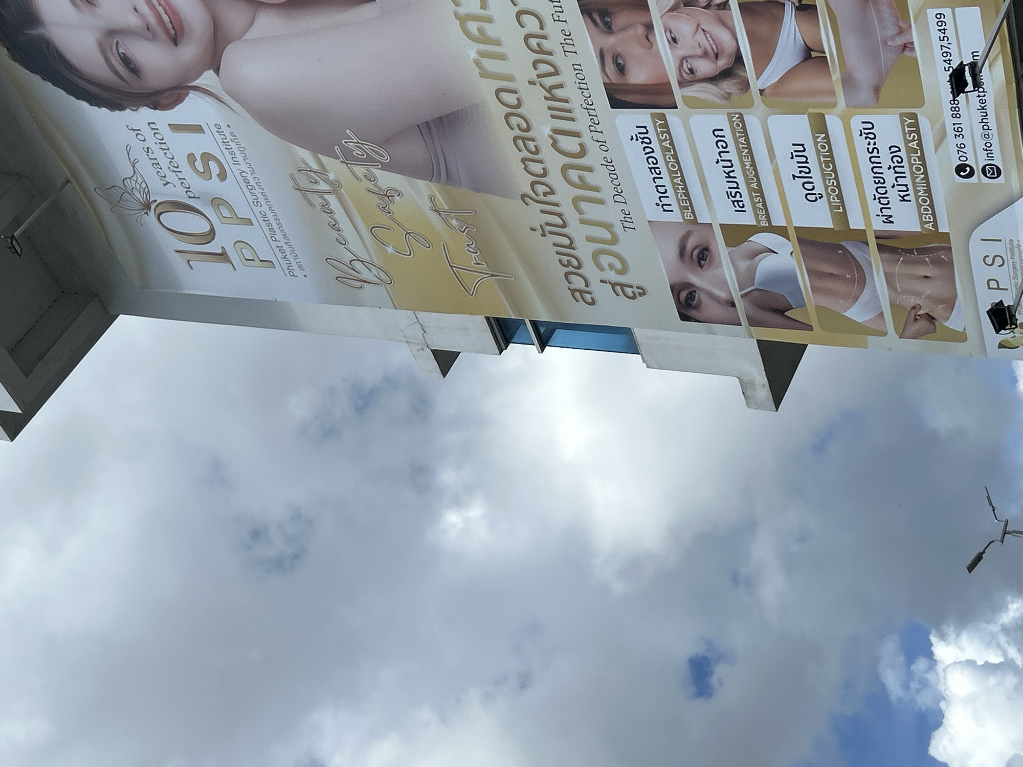 This is a close-up, eye-level, exterior shot of a large billboard advertising "Phuket Plastic Surgery Institute" (PPSI) in Wichit, Thailand. The billboard features images of women showcasing various cosmetic procedures. Text on the billboard includes "10 years of perfection," "Beauty," "Safety," "Trust," and "The Decade of Perfection The Future." Several medical procedures are listed in Thai and English, such as "BLEPHALOPLASTY" (eyelid surgery), "BREAST AUGMENTATION," "LIPOSUCTION," and "ABDOMINOPLASTY" (tummy tuck). Contact information, including a phone number "076 361 888" and email "info@phuketp...com," is also visible. The sky in the background is cloudy with some patches of blue. A street light is visible in the lower left portion of the frame.