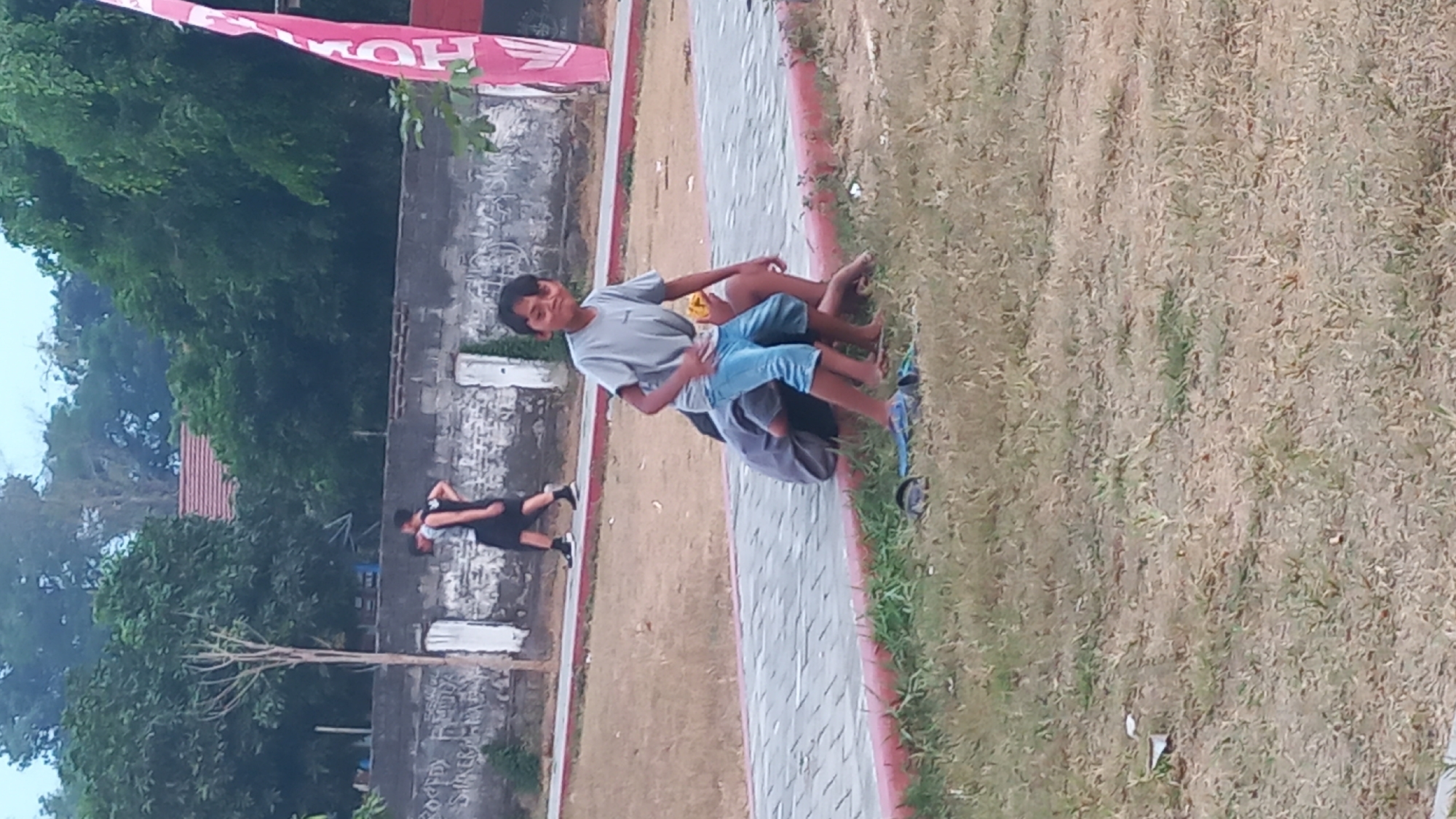 The image depicts an outdoor scene at the Pare City Park in Pare, Indonesia. In the foreground, a young boy in a grey t-shirt and blue shorts is standing and looking towards the camera, with a slight smile. He is positioned on a patch of dry grass. Behind him, another person, partially obscured, appears to be sitting on the ground, also on the grass. To the left of the central figures, a paved pathway with a red border is visible, with two pairs of flip-flops lying on the grass near its edge. In the background, two individuals are walking away from the camera on the paved pathway. One is wearing a black tank top and black shorts, and the other is in a grey tank top and black shorts. A low, weathered brick wall with graffiti runs along the back of the pathway, and beyond that, lush green trees and foliage dominate the upper portion of the image, with a hint of a building with a red roof visible through the trees. A tall, pink and white flag is positioned on the far right side of the frame. The overall atmosphere suggests a casual daytime activity in a public park.