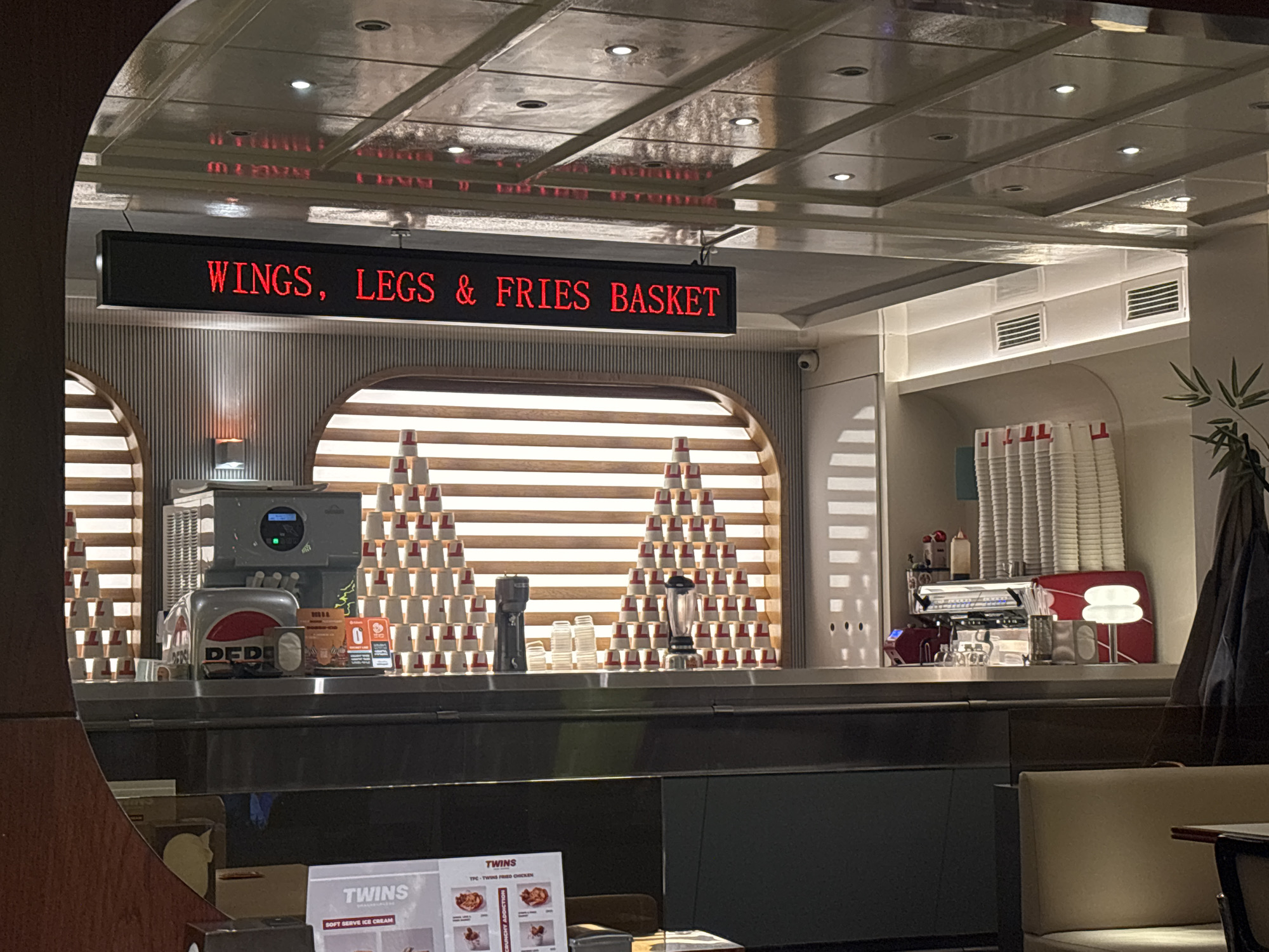 This image presents the interior of a modern, casual dining establishment, likely a restaurant or cafe, situated in Yerevan, Armenia. The scene centers on a well-organized service counter, brightly lit by overhead recessed lights embedded in a glossy, paneled ceiling that reflects a prominent LED sign.
The black LED sign, suspended above the counter, displays the text "WINGS, LEGS & FRIES BASKET" in illuminated red letters. Below it, the counter is equipped with various machinery and items. To the left, a soft-serve ice cream machine is visible next to a dispenser marked with the "DEDE" logo. Flanking these machines are intricate, pyramid-like arrangements of small paper cups, each with a red rim. Further to the right, a blender and a professional espresso machine with grinders are situated, accompanied by stacks of larger white cups. The background features decorative wall panels with horizontal slats and arched openings, contributing to a contemporary aesthetic. Vents are visible high on the back wall, and a potted plant adds a subtle touch of green on the far right.
A partially visible menu in the foreground confirms the establishment is named "TWINS," offering "SOFT SERVE ICE CREAM" and "TPC - TWINS FRIED CHICKEN." No people are present in the image, and the artificial lighting suggests an indoor setting, not indicating a specific time of day. The overall impression is of a clean, efficient, and inviting eatery.
