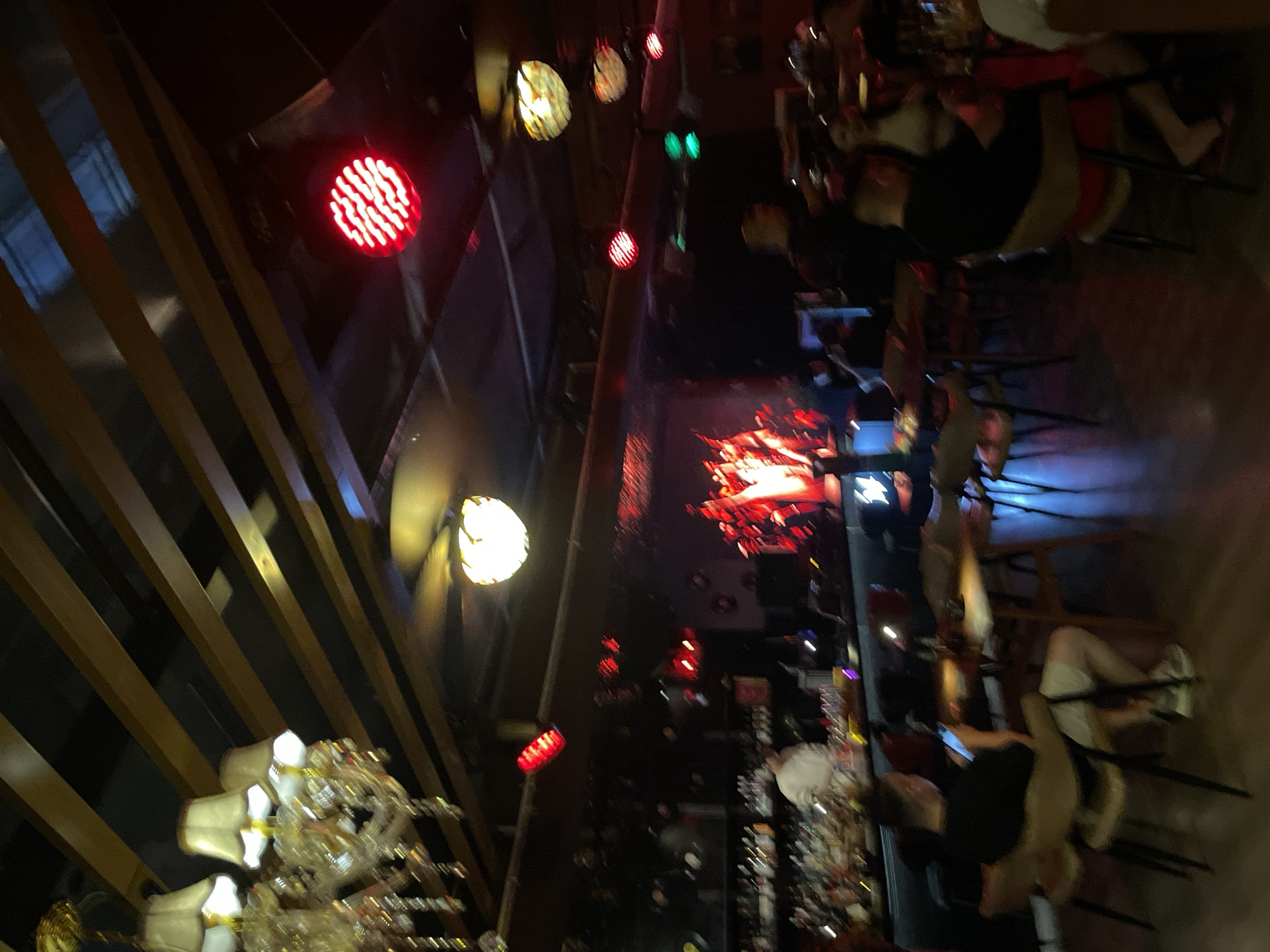 This image captures a dimly lit interior scene, likely a bar or music venue, in Liangjiang, China. The ceiling features exposed wooden beams and a large, ornate chandelier with multiple lamps. Several red, circular lights are visible, some illuminated and appearing to be stage or ambient lighting. In the background, a stage area is set up with a band performing, characterized by bright red and white stage lights. A bar area is visible with bottles and glassware. Several patrons are seated at tables and at the bar, with one individual in the foreground looking at their phone. The overall atmosphere suggests a lively evening at an entertainment establishment.
