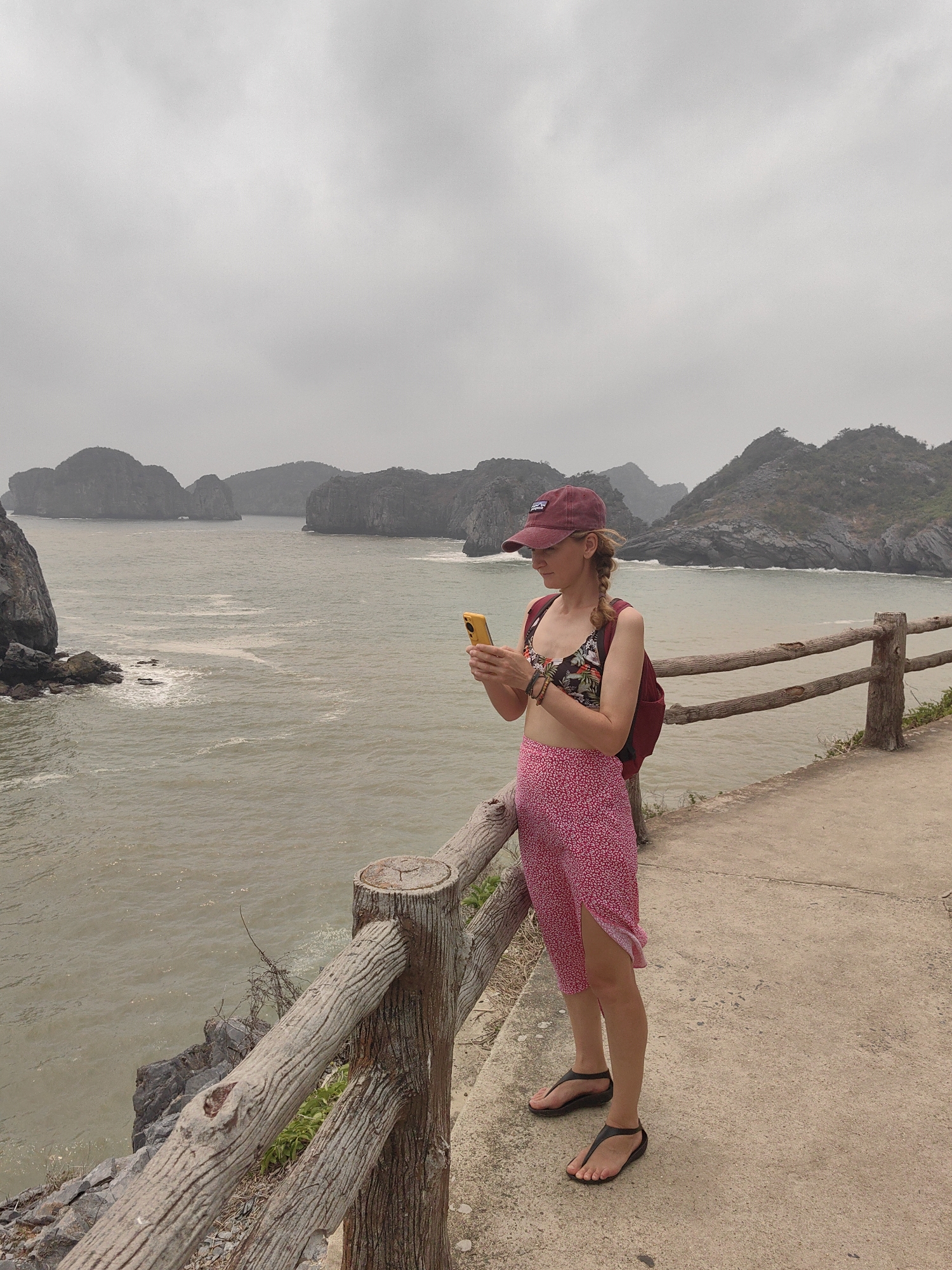 A woman stands on a concrete path beside a wooden railing, looking at her yellow smartphone. She wears a maroon baseball cap, a floral print top, a red backpack, a red and white floral skirt, and black sandals. In the background is the ocean, and a rocky coastline with islands under a cloudy sky. The scene is likely taken in the city of Hải Phòng, Vietnam.
