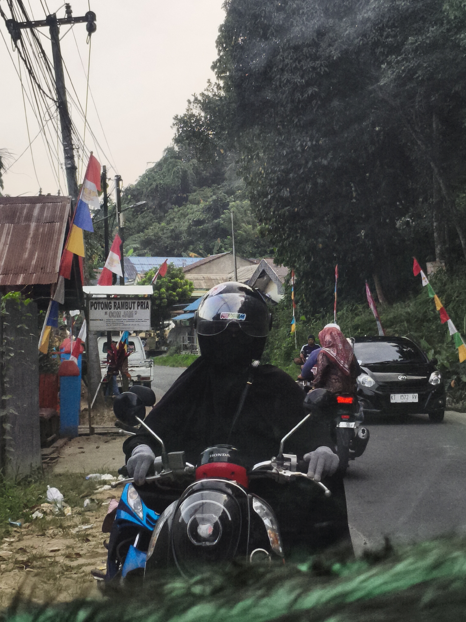 A motorcycle rider, wearing a black abaya and helmet, is on a road in Samarinda, Indonesia. The rider's hands, covered in gray gloves, are on the handlebars of a black scooter. The scooter is blue and black. Behind the rider, a black car with the license plate "KT 1572 RV" is driving on the road. Another motorcycle is visible behind the car, with two people wearing hijabs. The surroundings show a roadside environment with trees, houses, and flags. A sign above a shop reads "POTONG RAMBUT PRIA" (Men's Haircut). The sky is overcast.