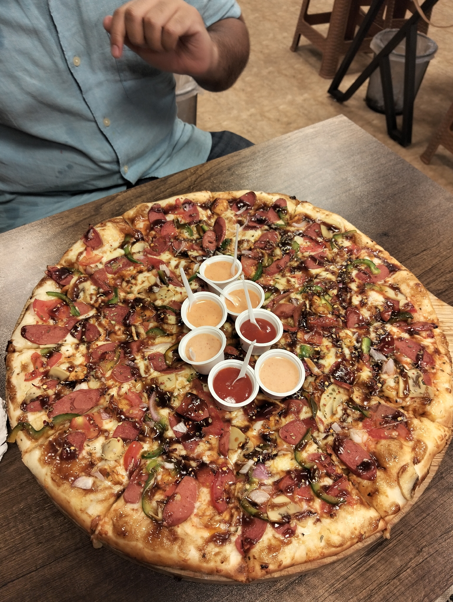 A slightly high-angle shot depicts a large pizza on a wooden table, likely indoors. The pizza is topped with sausage slices, green peppers, tomatoes, onions, mushrooms, and a dark sauce drizzled over the ingredients. Seven small plastic cups filled with sauces of varying orange and red hues are placed in the center of the pizza, each with a small white spoon inserted. A person wearing a light blue shirt is seated at the table, with their hand visible in the upper left corner. In the background, there is a wastebasket and several stools with woven seats and black metal frames. Based on the available details, it's difficult to confirm its location in Dhaka, Bangladesh with certainty.