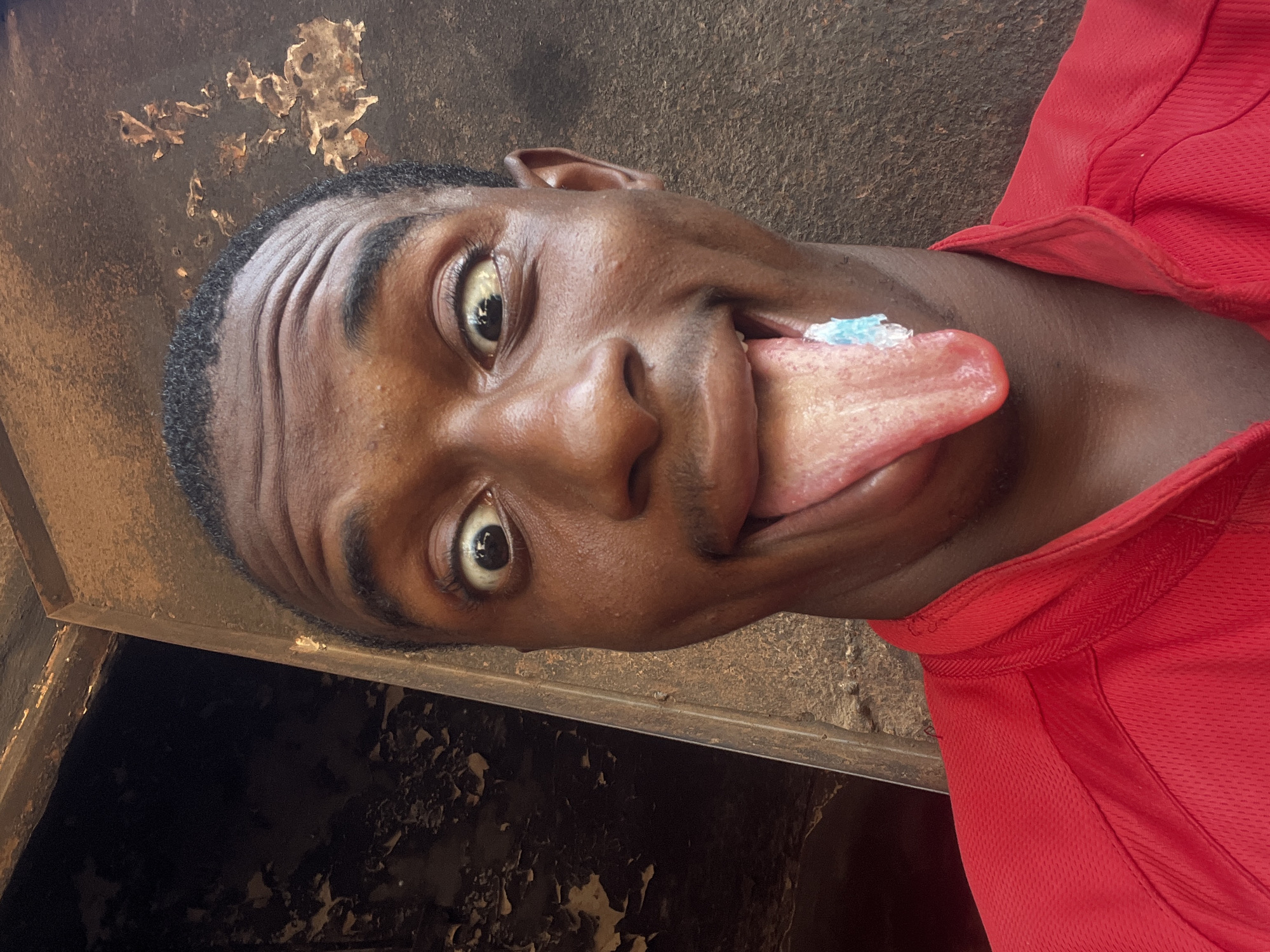 A selfie-style, close-up shot captures a young Black man in a red mesh shirt. His head is tilted back, eyes wide, and tongue extended with a blue substance on the tip. The background consists of a dark, textured surface, appearing to be a worn or peeling wall or structure. Due to the angle and lack of clear landmarks, identifying the specific location in Onitsha, Nigeria, is not possible from the image alone. No specific actions or interactions can be determined. The environment is only visible as a close-up of a weathered surface.