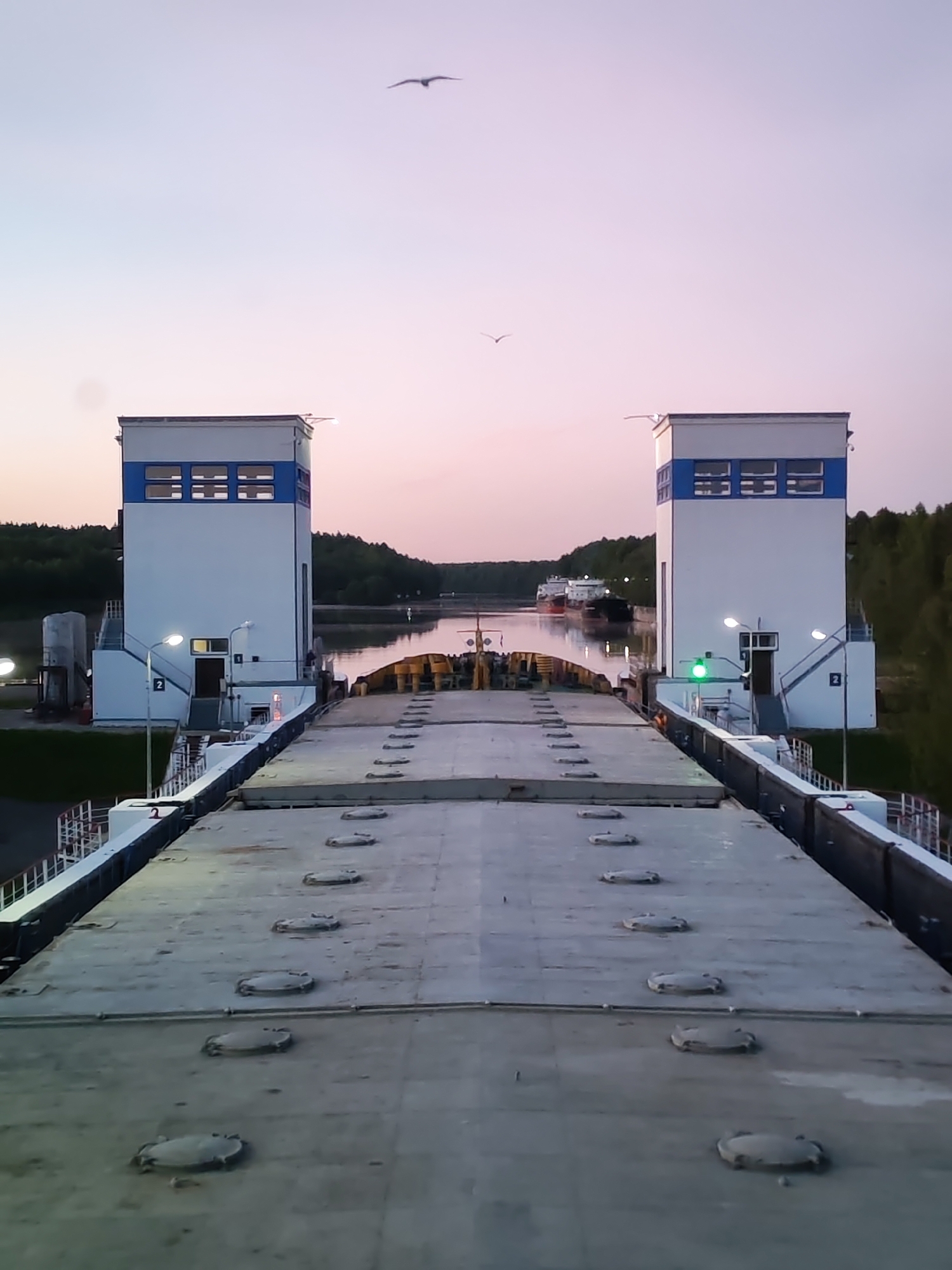 The image captures a tranquil scene of a canal lock or waterway system during twilight, likely dusk. The perspective is from the deck of a vessel, possibly a cargo ship or barge, looking forward into the passage.
In the foreground, the flat, gray deck of the vessel is visible, featuring a series of circular metal hatches or covers running down its length. A white railing marks the edge of the deck on the left.
The midground is dominated by two identical, tall, white buildings flanking the waterway. Each building features a distinctive blue horizontal stripe across its upper section and multiple rectangular windows. These structures appear to be control towers or operational buildings for the lock. Artificial lights illuminate the buildings and their surroundings, with a prominent green signal light visible on the right-hand building, potentially indicating permission to proceed. On the left building, the numbers "1" and "2" are visible near entranceways.
Beyond the lock, a calm body of water extends into the distance, reflecting the soft, gradient colors of the twilight sky—a blend of light blue, pink, and subtle purple hues. In the far distance, at least two large cargo ships or tankers are visible on the water, with dense green trees lining the banks of the canal.
Two birds are captured in flight against the sky; one is higher and more centrally located, while a smaller, fainter bird appears further to the left. The scene evokes a sense of calm and operational readiness amidst significant industrial infrastructure.