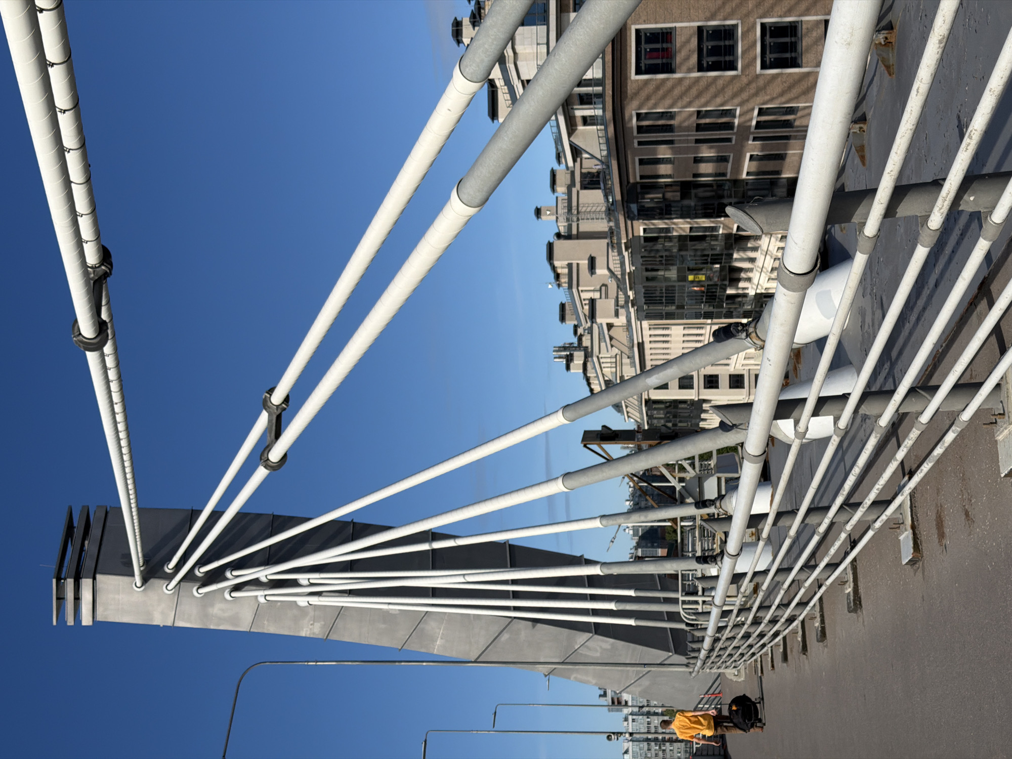 The image depicts a pedestrian bridge in Saint Petersburg, Russia, characterized by its modern cable-stayed design. A lone individual, wearing a bright orange shirt, walks away from the camera, pulling a black rolling suitcase. They are on the bridge walkway, which is bordered by a white metal railing. In the background, the cityscape of Saint Petersburg is visible, featuring contemporary apartment buildings with light-colored facades and dark-framed windows, some with visible red accents. The sky is a clear, vibrant blue, suggesting a sunny day. The bridge's main structural elements, including a large concrete pylon and numerous white cables, are prominent in the foreground, emphasizing the architectural style of the infrastructure.