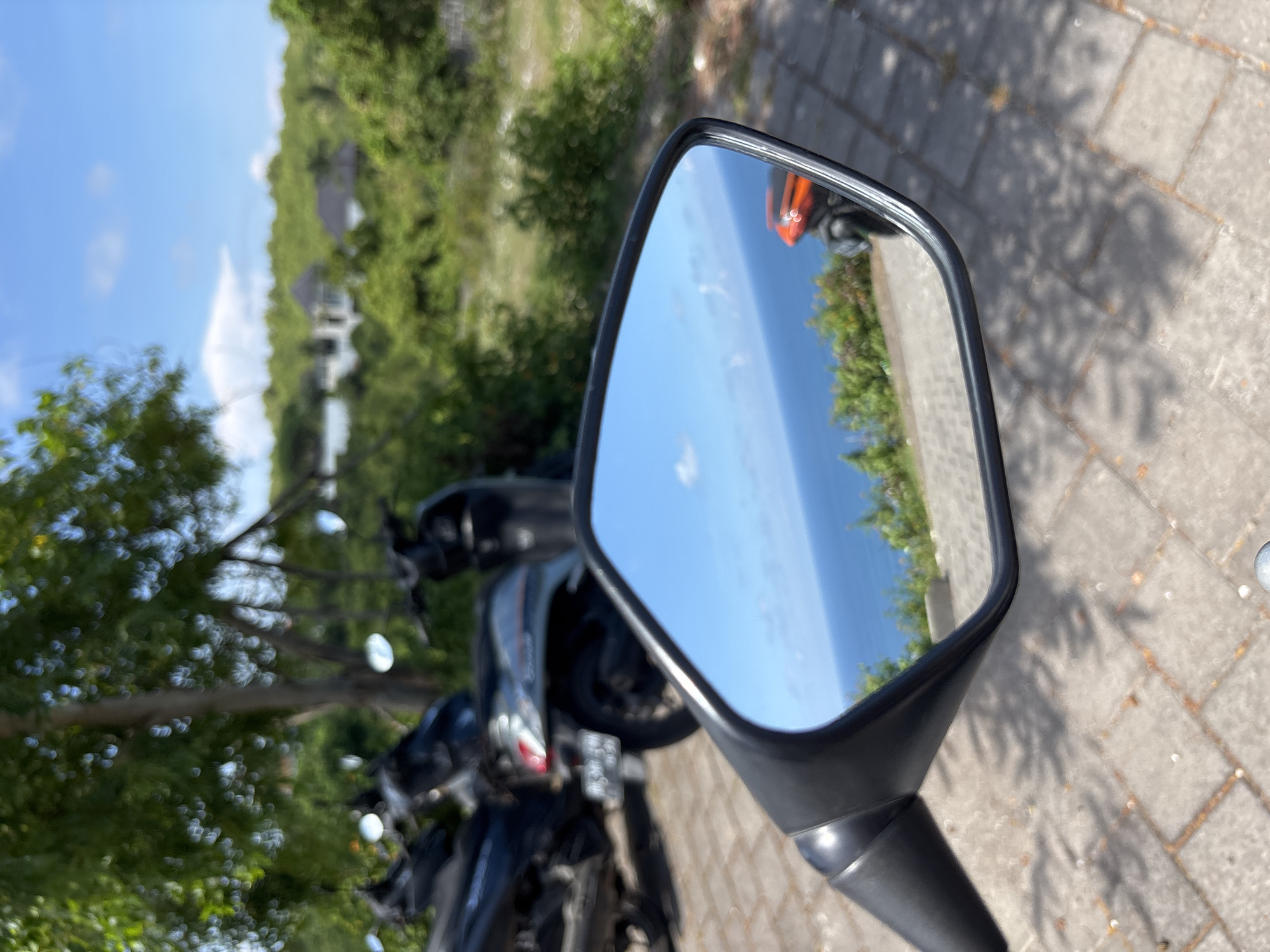 A motorcycle's side mirror reflects a coastal scene, suggesting a location near the ocean. The sky is clear and blue, reflecting in the mirror. Green foliage lines the coast. In the background, more greenery and a white building with a dark roof are visible. The motorcycle is parked on a paved surface. Based on the location metadata, the scene is near Ungasan, Indonesia.