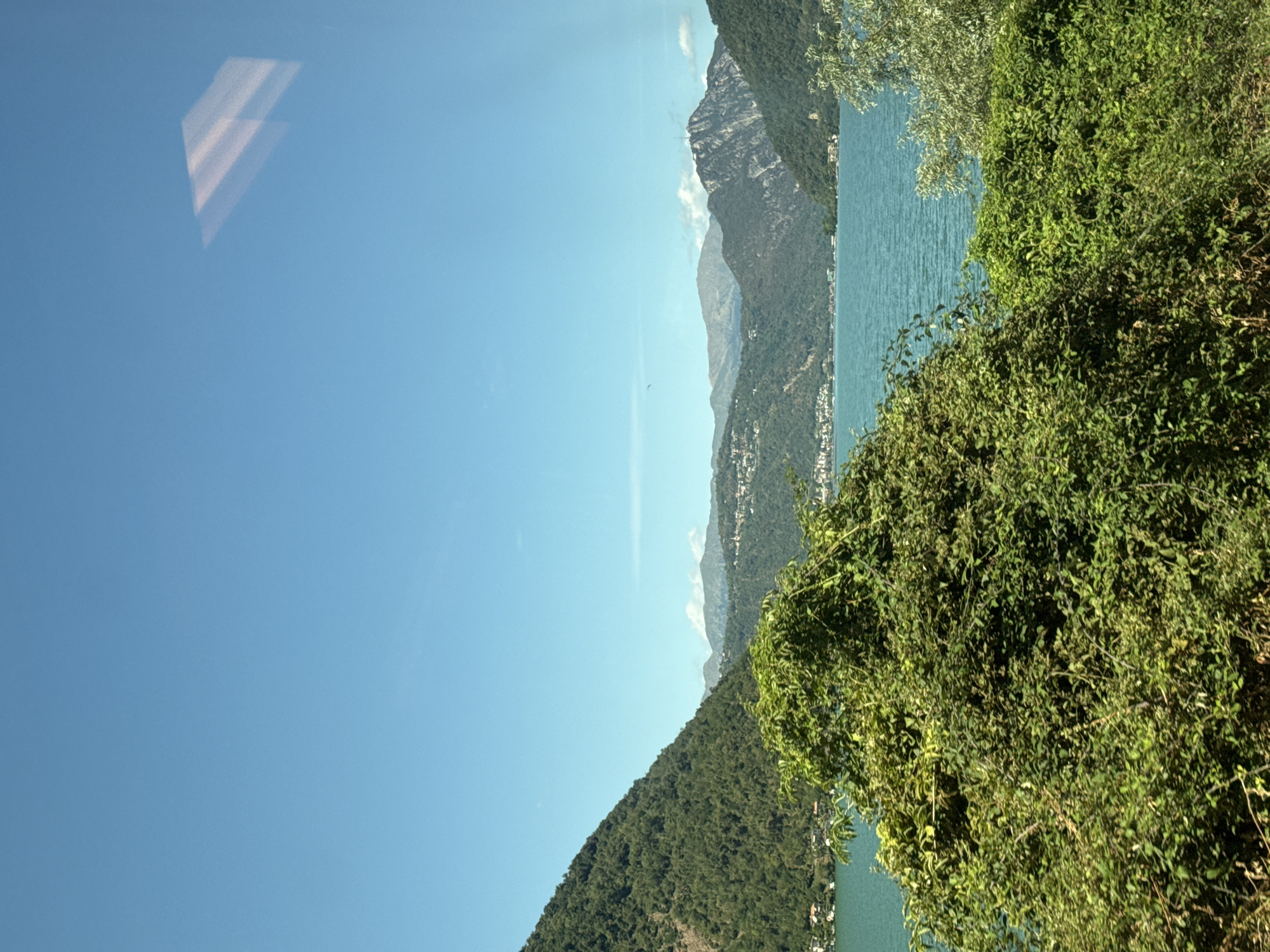 The image shows a scenic view of a body of water surrounded by lush green mountains. The water is a vibrant turquoise color, reflecting the clear blue sky. The mountains are covered in dense forests, with some buildings scattered along their slopes. In the foreground, there is a thick patch of green foliage, obscuring the view of the lower part of the scene. The sky is clear with a faint reflection visible in the top right corner, possibly from a window. The overall impression is one of natural beauty and tranquility. The location is likely Lugano, Switzerland, specifically the area around the lake, which is a popular tourist destination known for its stunning alpine scenery and pristine waters. The image does not contain any people or specific objects that indicate a precise location within the city.