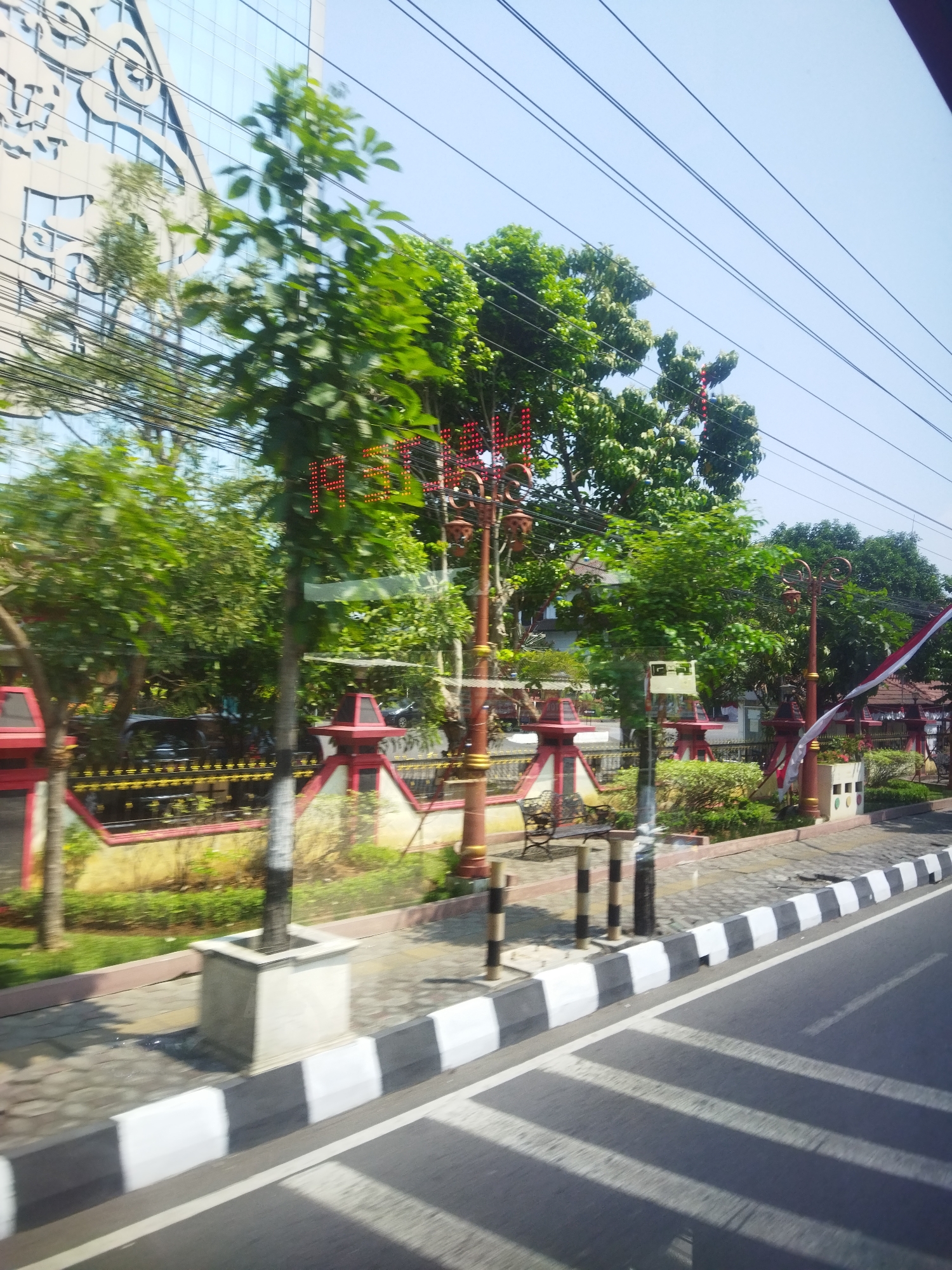 The image depicts a street scene in Jombor, Indonesia, on a clear day with a bright blue sky. The foreground shows a paved road with white and black striped speed bumps and a curb. Beyond the curb, a sidewalk is visible, with a small square concrete planter containing a slender tree trunk. Several decorative street lamps with red accents and light fixtures are present along the sidewalk.
Behind the sidewalk, there is a low wall with ornate yellow and black detailing. Green grass and bushes are planted behind the wall. Further back, a building with a modern, reflective facade can be seen, with large windows and decorative elements. Several trees with lush green foliage are interspersed throughout the scene, some obscuring parts of the buildings and sky. In the middle ground, there is a red and white striped flag or banner hanging from a pole. A bench is visible on the sidewalk. Red illuminated text, appearing to spell "P" and "F" followed by other unidentifiable letters, is mounted on a pole. A few cars are parked behind the decorative wall. The image appears to be taken from a moving vehicle, as indicated by some motion blur.