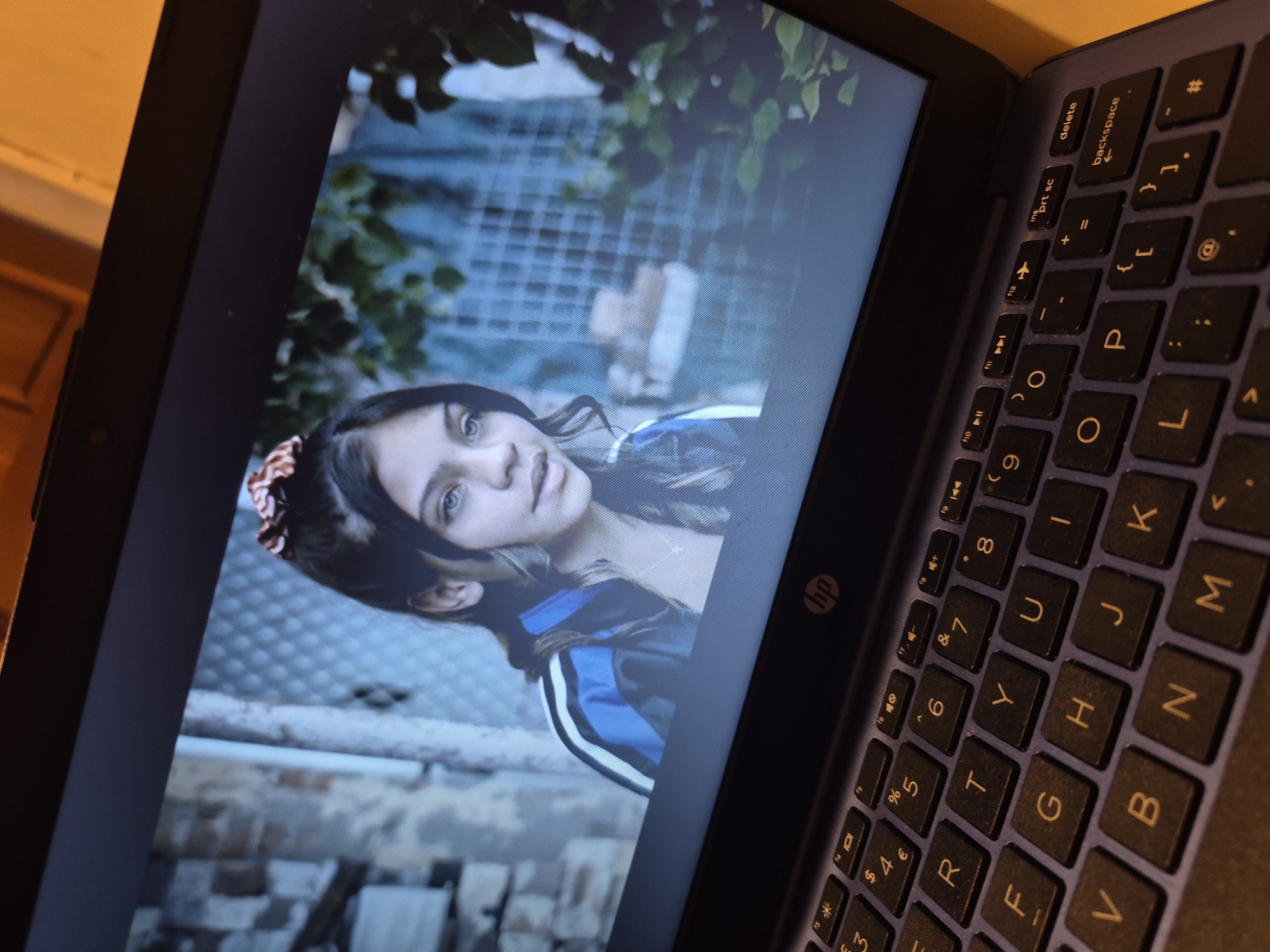 The image shows a laptop screen displaying a scene featuring a young girl with striking blue eyes and dark hair tied up with a scrunchie. She is wearing a blue and white jacket and a necklace. The background is slightly blurred, showing what appears to be a brick wall and some foliage. The laptop itself is open and in focus, with a partial view of its keyboard visible in the foreground. The keyboard has the HP logo on it. The lighting suggests an indoor setting with ambient light possibly coming from the screen itself. There is no discernible information about a specific location in Sandwell, United Kingdom within the image.