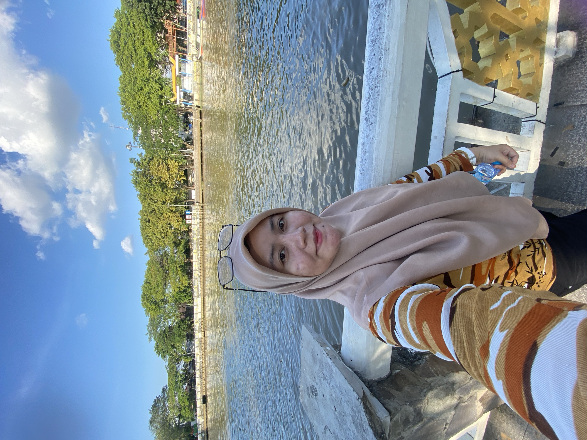 A woman is taking a selfie on a bright, sunny day. She is wearing a beige hijab and a long-sleeved shirt with an orange, brown, and white camouflage pattern. She is holding a clear plastic bottle of water in her right hand. She is standing on a paved walkway next to a body of water, with trees and buildings visible on the opposite bank. In the background, there are boats docked along the shore. The scene is likely the Martapura River in Banjarmasin, Indonesia, a city known for its extensive river system and floating markets.