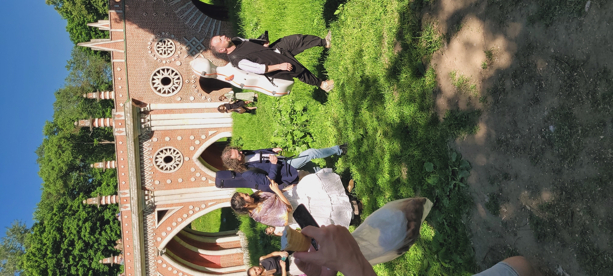 The image depicts an outdoor scene at the Tsaritsyno Museum-Reserve in Moscow, Russia, specifically featuring the distinctive red brick architecture of the Figured Bridge (or Grand Bridge) in the background. The bridge exhibits multiple Gothic Revival-style arches, circular decorative windows with radial spoke patterns, and ornate finials along its upper edge. Lush green trees are visible behind and above the bridge under a clear blue sky.
In the midground, a group of people is walking across a grassy area with some patches of dirt. From left to right:
1. Partially visible hands in the extreme foreground hold an ice cream cone and a black smartphone, seemingly recording or photographing the scene.
2. Below the hands, a clear plastic bag filled with brown, lumpy objects (possibly nuts or small snacks) is present.
3. A woman, possibly seated or crouching, wears a white dress and a multi-patterned shawl. Her dark hair is long, and she appears to be looking towards the man with the guitar case.
4. A man with long, curly light brown hair, wearing a blue jacket over a white shirt, light blue jeans, and sunglasses, walks towards the right. He carries a dark blue soft guitar case on his back and holds a green water bottle.
5. A man with short dark hair and a beard walks further to the right, wearing a black sleeveless tunic over a white long-sleeved shirt, black pants, and light-colored sneakers. He carries a large, light-colored (likely white or cream) hard cello case on his back.
In the background, beyond these individuals but in the grassy area, two women are walking: one in a black dress with a handbag, and another in a light-colored patterned dress. Indistinct figures are also visible on top of the Figured Bridge. The scene is brightly lit by sunlight, casting shadows on the grassy ground.