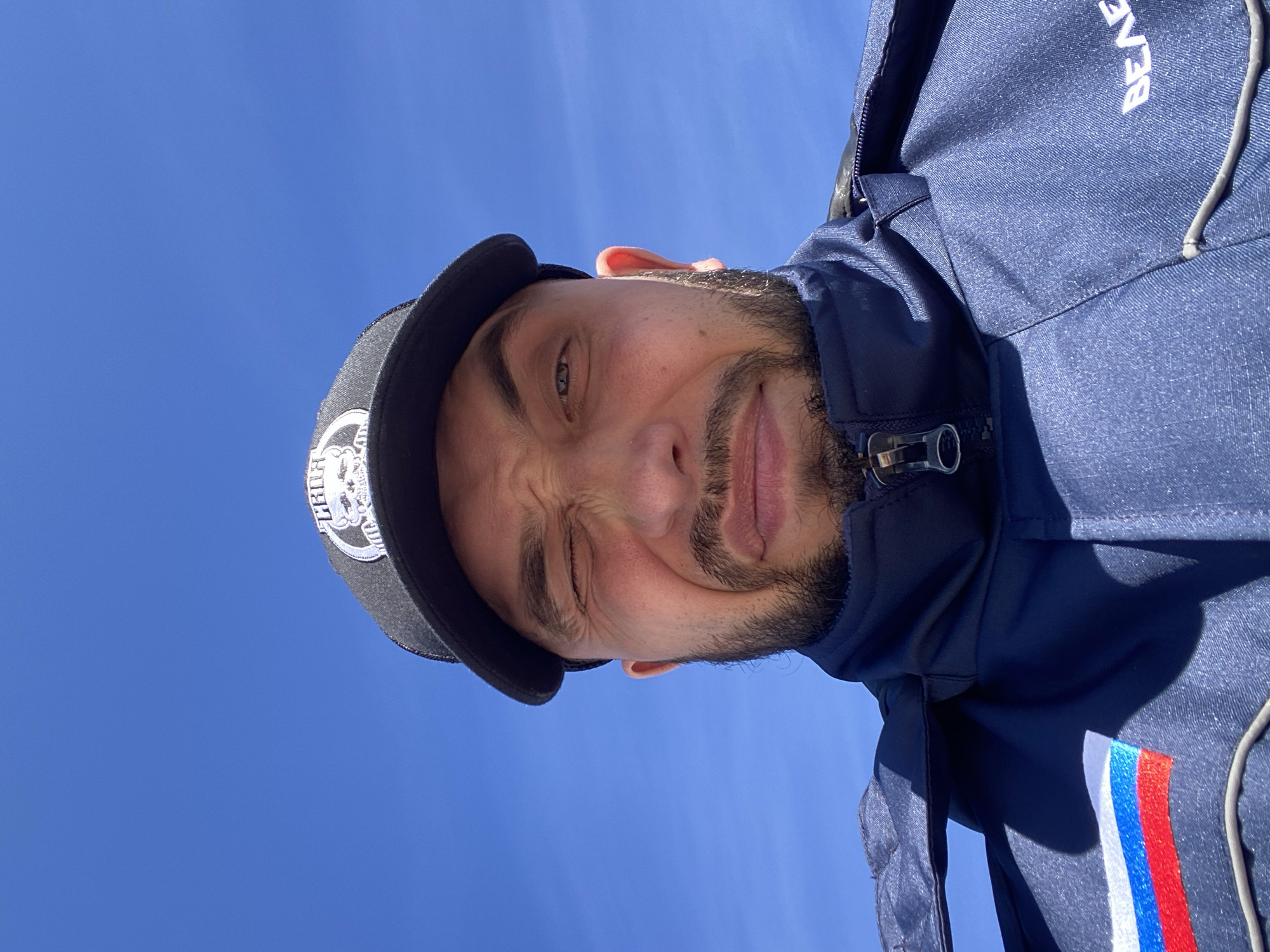A low-angle shot captures a man's head and upper torso against a clear blue sky. The image is rotated 90 degrees counter-clockwise. The man looks towards the viewer with a playful expression, winking with his right eye. He has a beard and mustache. He is wearing a black baseball cap with white embroidery, part of which shows the Cyrillic text "КНЯЗ" (KNYAZ). He is dressed in a dark blue, possibly athletic or outdoor, jacket with a zipper. On the upper right shoulder area of the jacket, the white text "BEAR" is visible, matching the image's rotation. A prominent vertical striped emblem in white, blue, and red, representing the colors of the Russian flag, is visible on the right side of his jacket. The background is an unblemished, bright blue sky, suggesting sunny and clear weather during the daytime. The man's expression conveys a lighthearted and casual mood. The location context for the image is Luzhitsy, Russia, aligning with the national colors displayed on his attire.