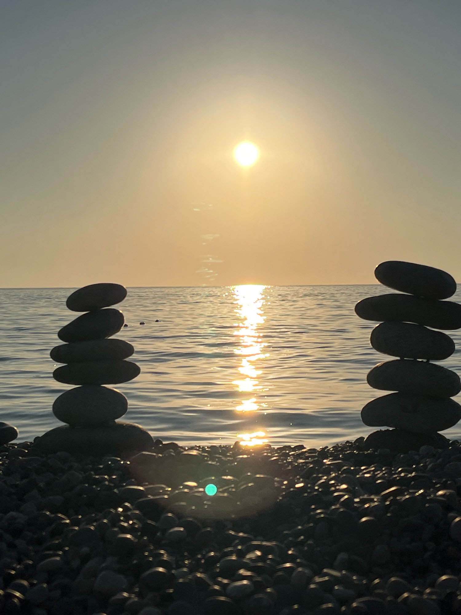 The image depicts a tranquil beach scene at sunset in Kobuleti, Georgia. The sun, a bright orb, is low on the horizon, casting a golden glow across the water and sky. The sea is calm, with gentle ripples reflecting the sunlight. Two stacks of smooth, dark stones are balanced on the pebbled shore. One stack stands on the left, composed of five stones, while the other, on the right, is made of six stones. The foreground is filled with a dense scattering of pebbles, also dark in color and catching the fading light. The overall impression is one of serenity and natural beauty.