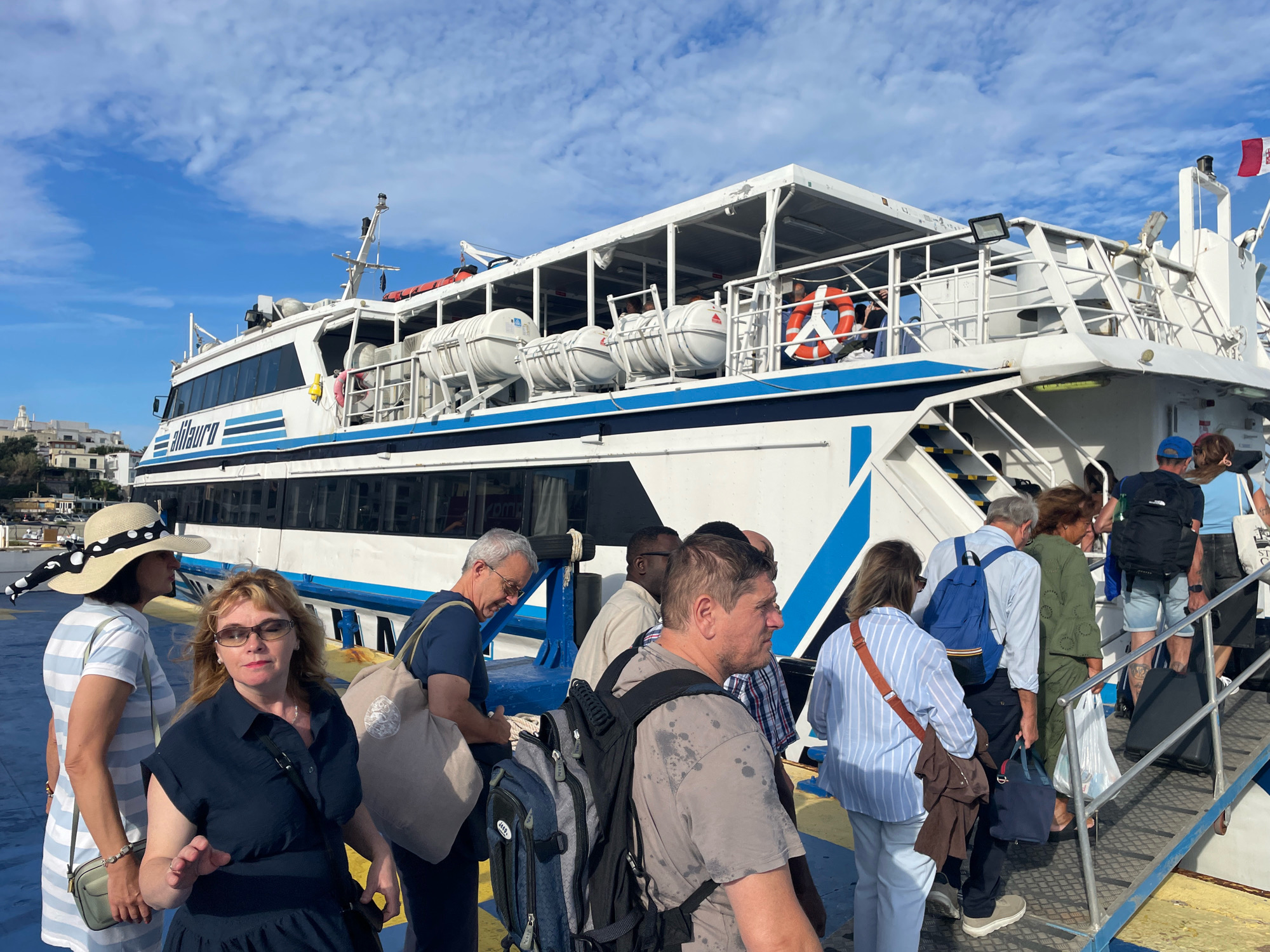The image shows a group of people boarding a ferry in Forio, Italy. The ferry is white with blue trim and has the name "Alilauro" written on its side. The people are dressed casually and are carrying bags and backpacks. The sky is blue with some clouds. The scene appears to be a sunny day at a port.