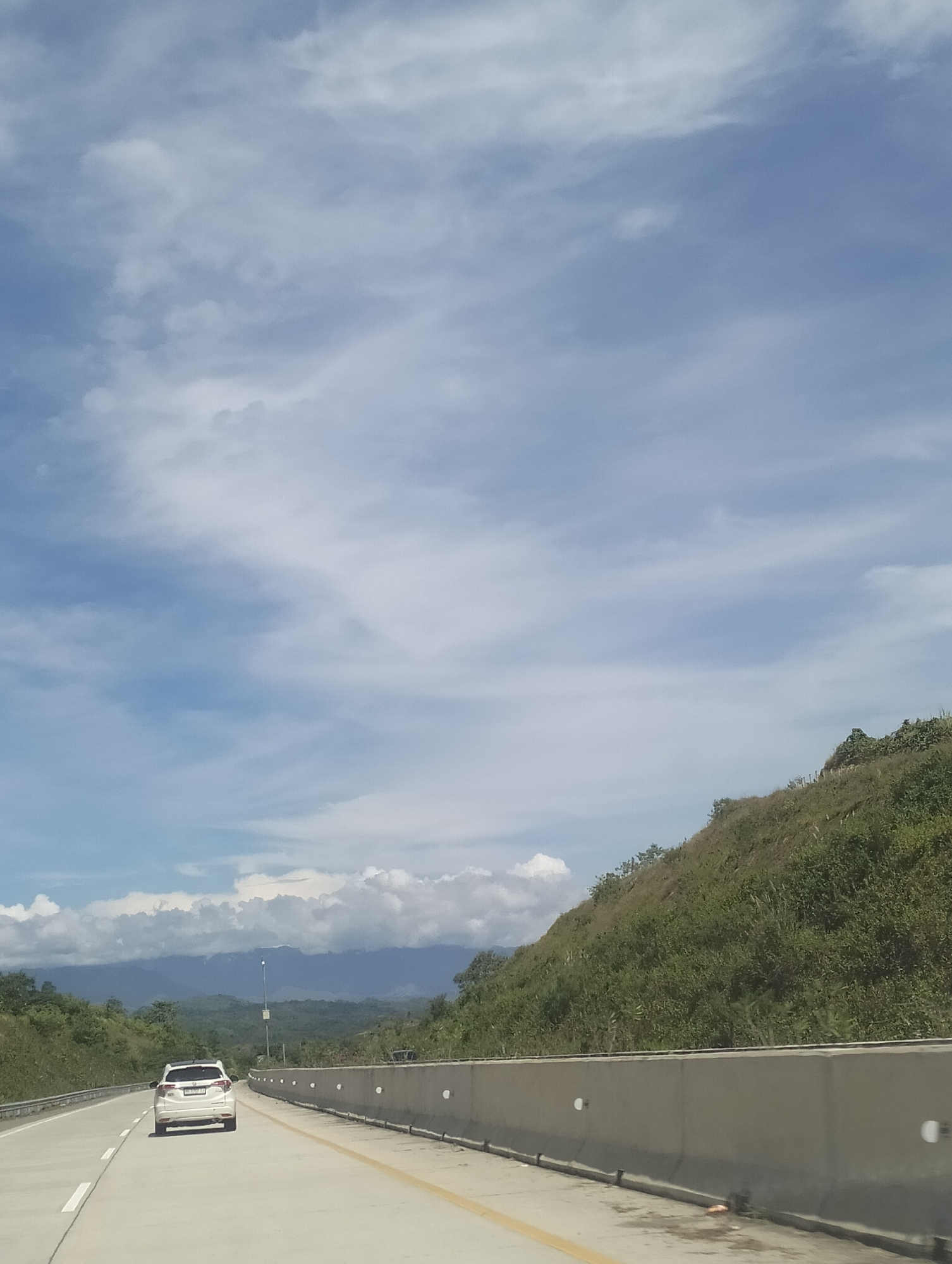 The image depicts a scenic view from a highway in Lamtamot, Indonesia. A white SUV is traveling on the right lane of the road, heading away from the viewer. The highway has two lanes in each direction, separated by a yellow line and white lines. On the right side of the highway, there is a concrete barrier, and beyond that, a grassy hill covered in lush vegetation. The background features rolling hills and mountains, shrouded in mist under a partly cloudy sky. The clouds are white and wispy, scattered across a bright blue sky. The overall impression is one of a peaceful journey through a natural landscape.