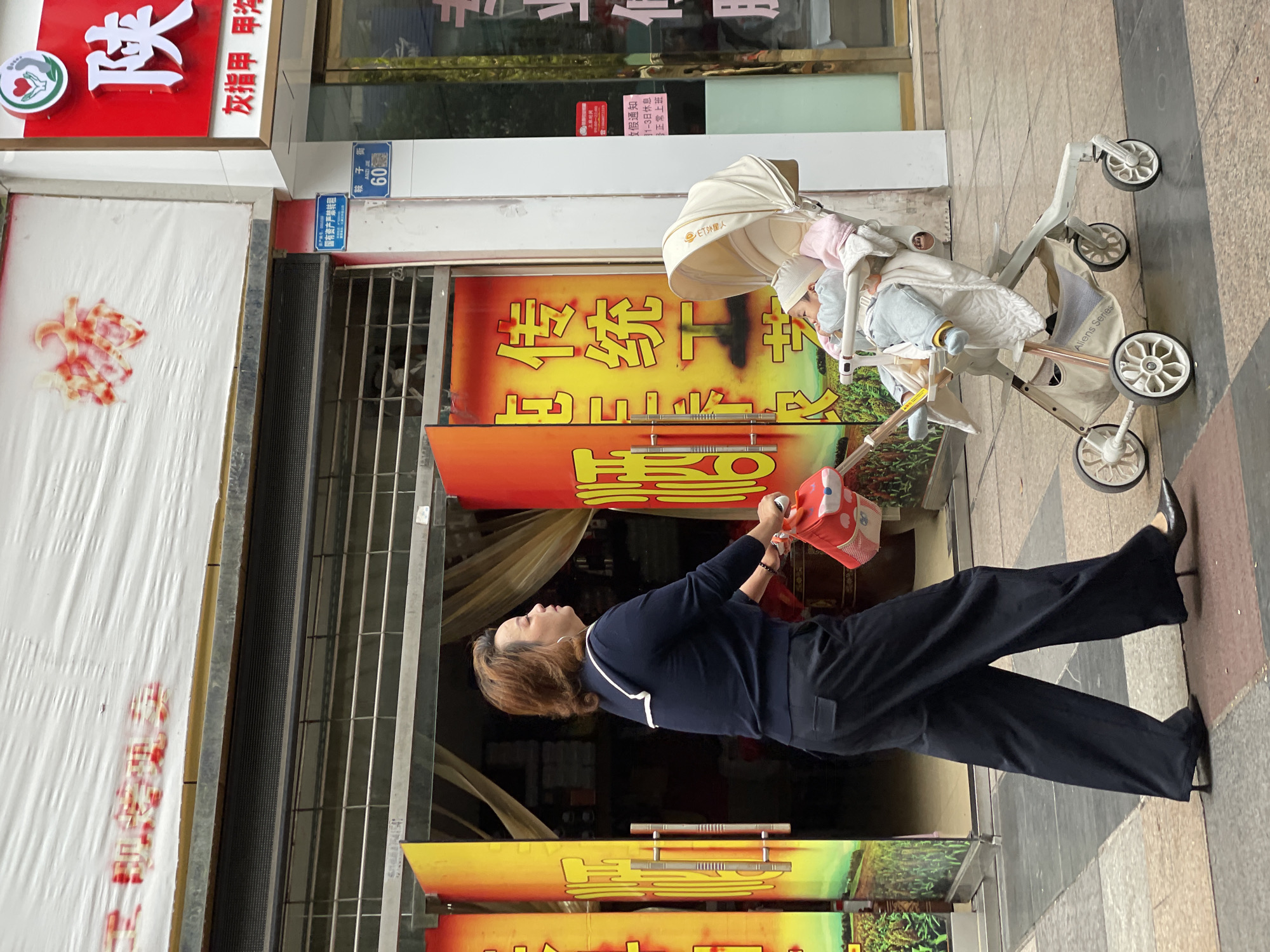 A medium shot shows a woman standing next to a baby in a stroller outside a business in Jiangjin, China. The woman is wearing black pants, a dark blue top with white trim, and black heels. She is holding a red bag with white trim. The baby is lying in a white stroller with the canopy up. The building behind them is two stories and has a yellow and orange sign with Chinese characters on it. Next to the sign is the address: Anzi Street, Number 60. A sign next to that says, "State-owned assets are strictly prohibited from being transferred."